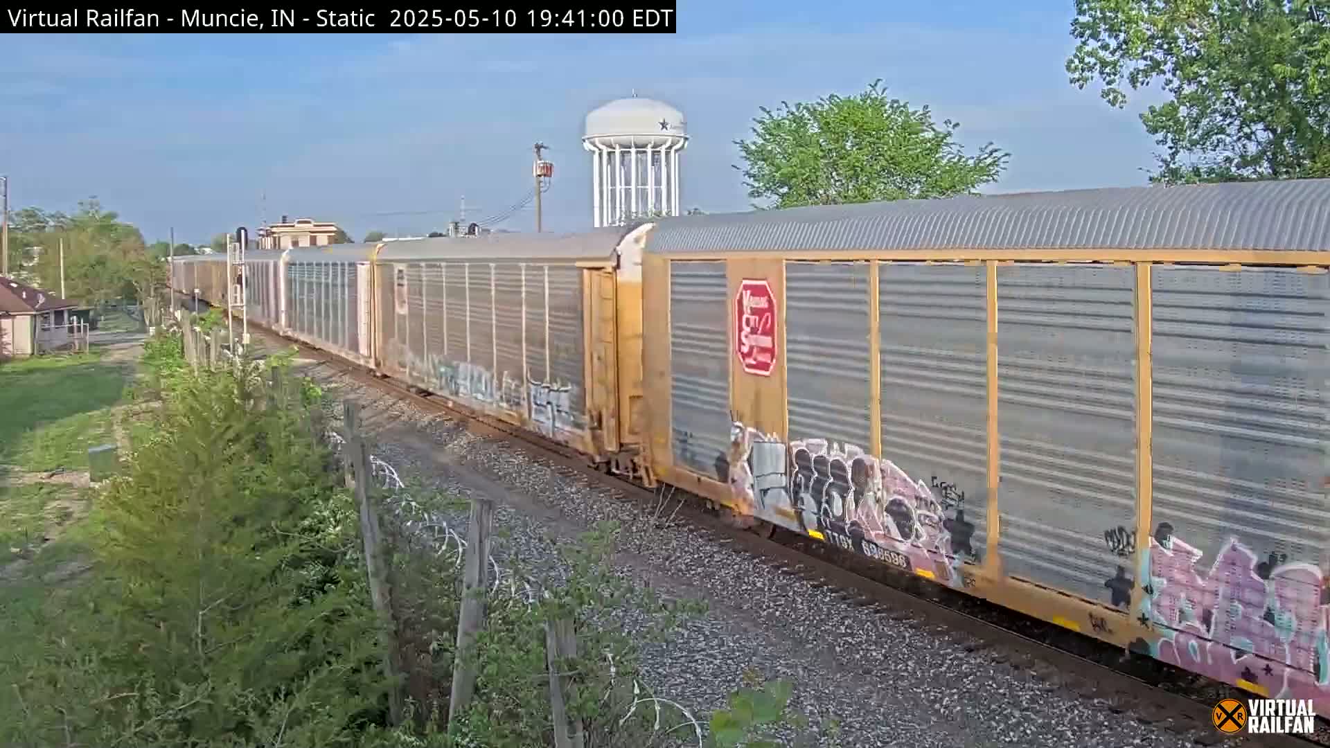A long freight train passes by under a clear blue sky, near a water tower and some trees.