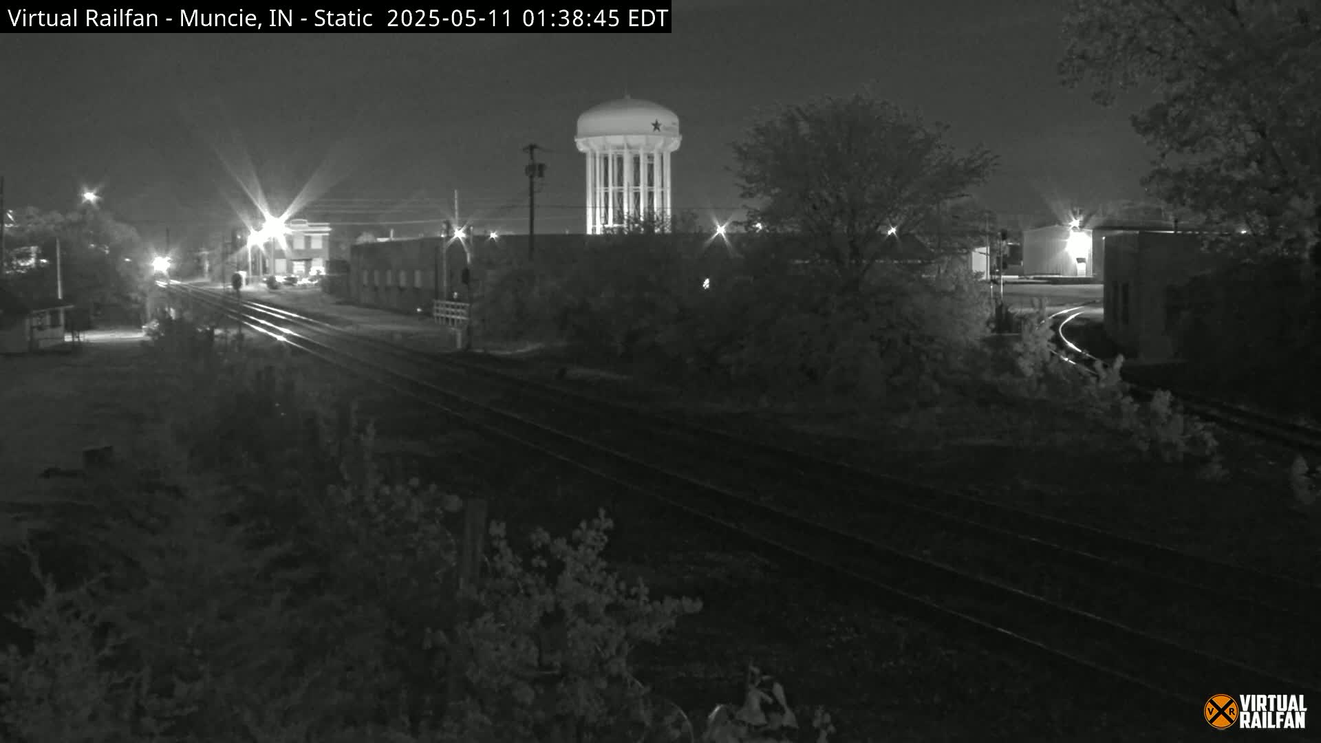 A nighttime view of railroad tracks leading toward a water tower and industrial buildings under a clear, dark sky.