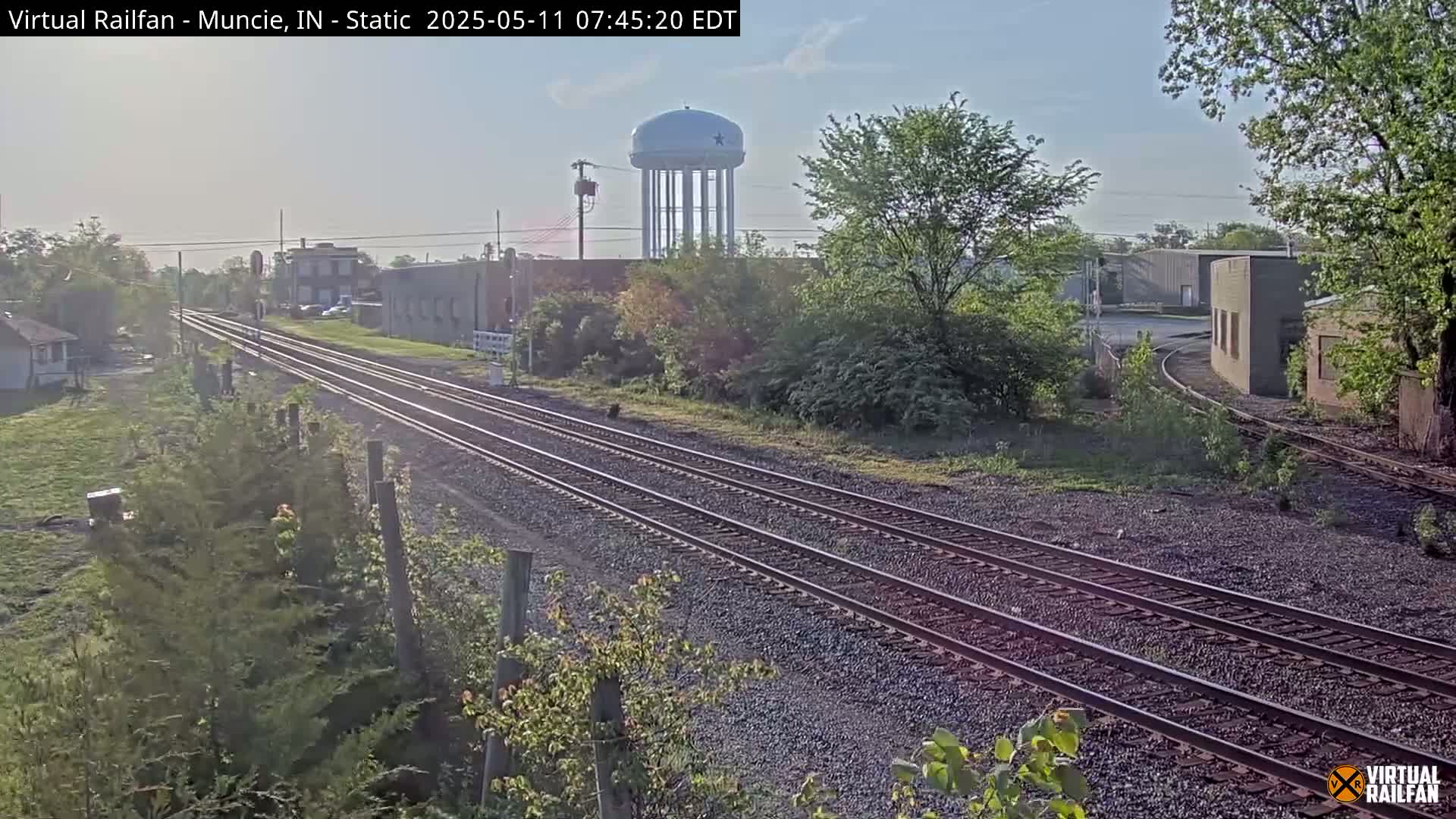 A clear, sunny day reveals multiple train tracks curving through a landscape of low buildings, vegetation, and a water tower.