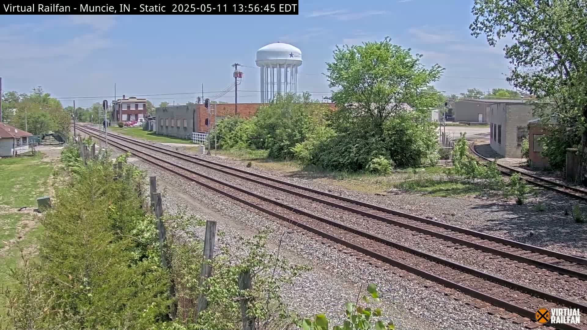 A sunny view of railroad tracks extending into the distance, passing industrial buildings and a water tower, surrounded by greenery.