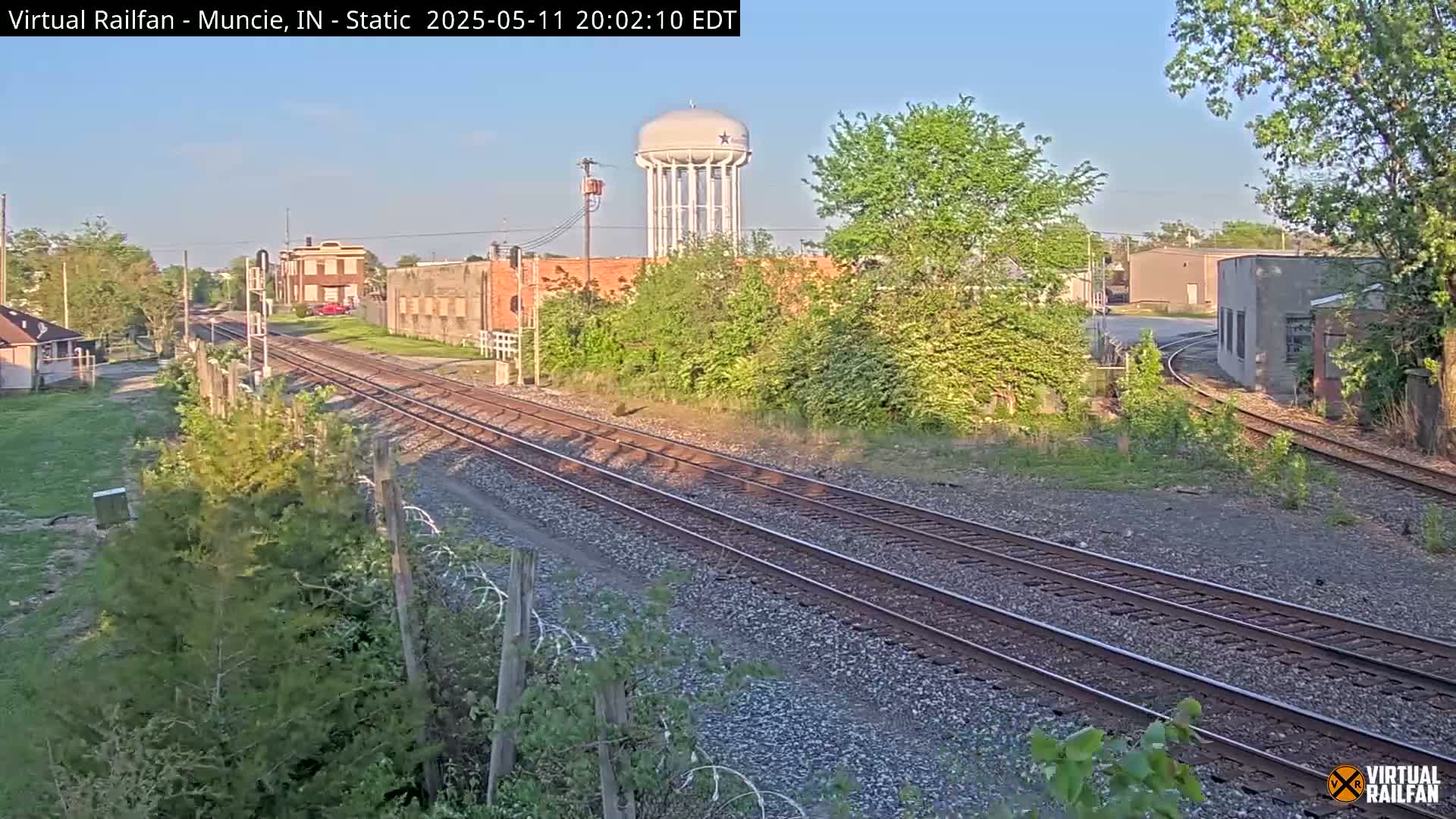 Under a clear evening sky, railroad tracks extend into the distance past industrial buildings and a water tower, surrounded by greenery.
