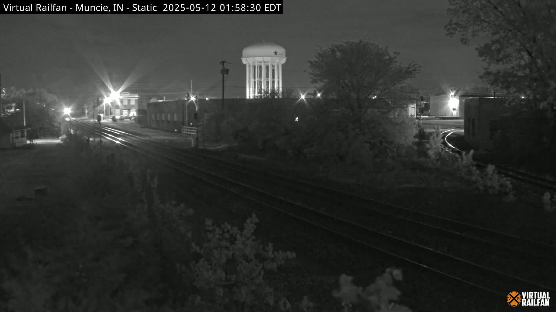 A nighttime, low-light view of railroad tracks leading toward a water tower and industrial buildings, surrounded by trees.