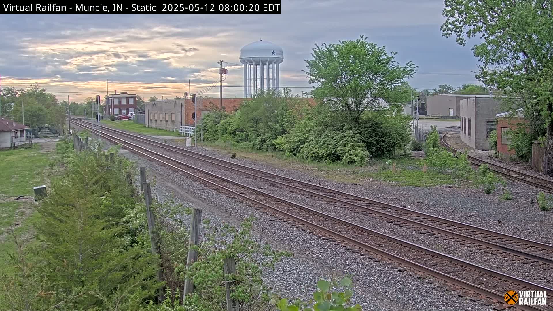 Two sets of train tracks extend into the distance past industrial buildings and a water tower under an overcast sky.