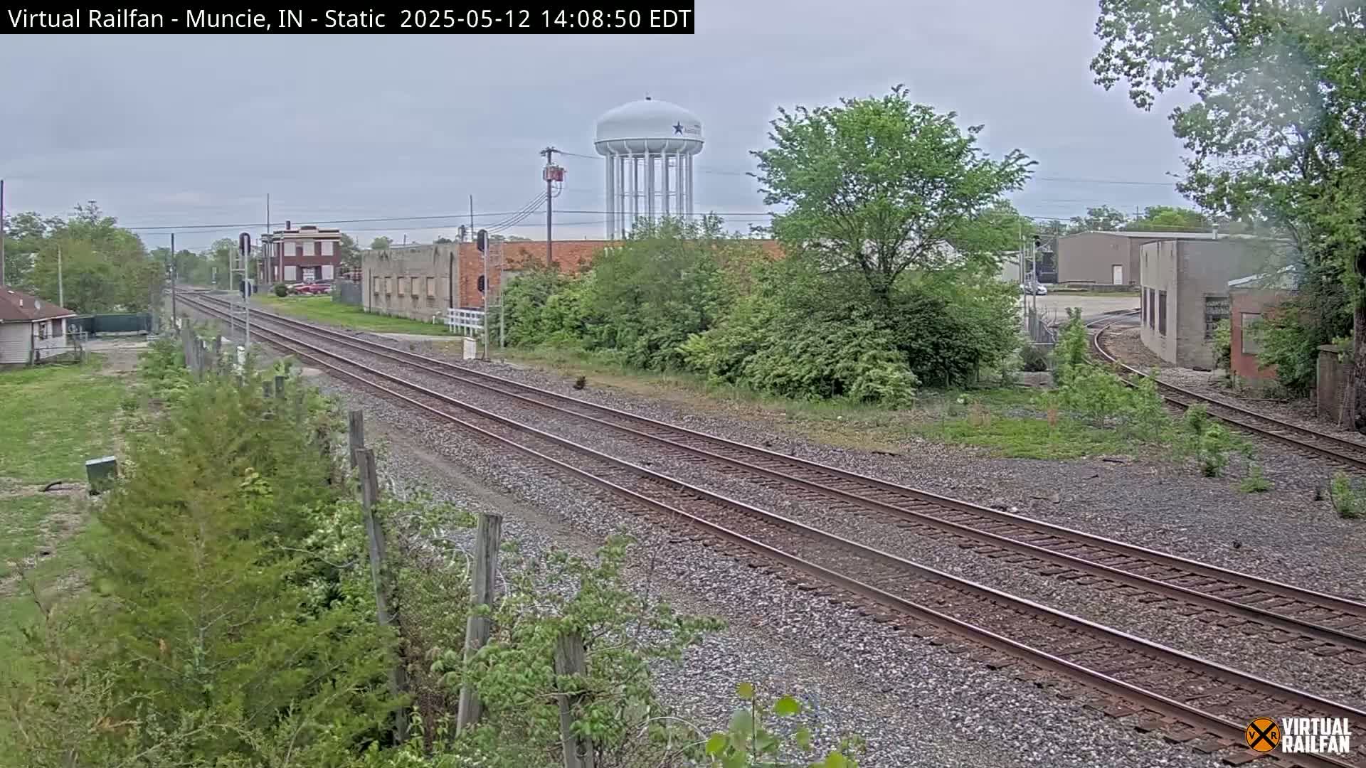 A view of railroad tracks extending into the distance, passing industrial buildings and a water tower under an overcast sky.