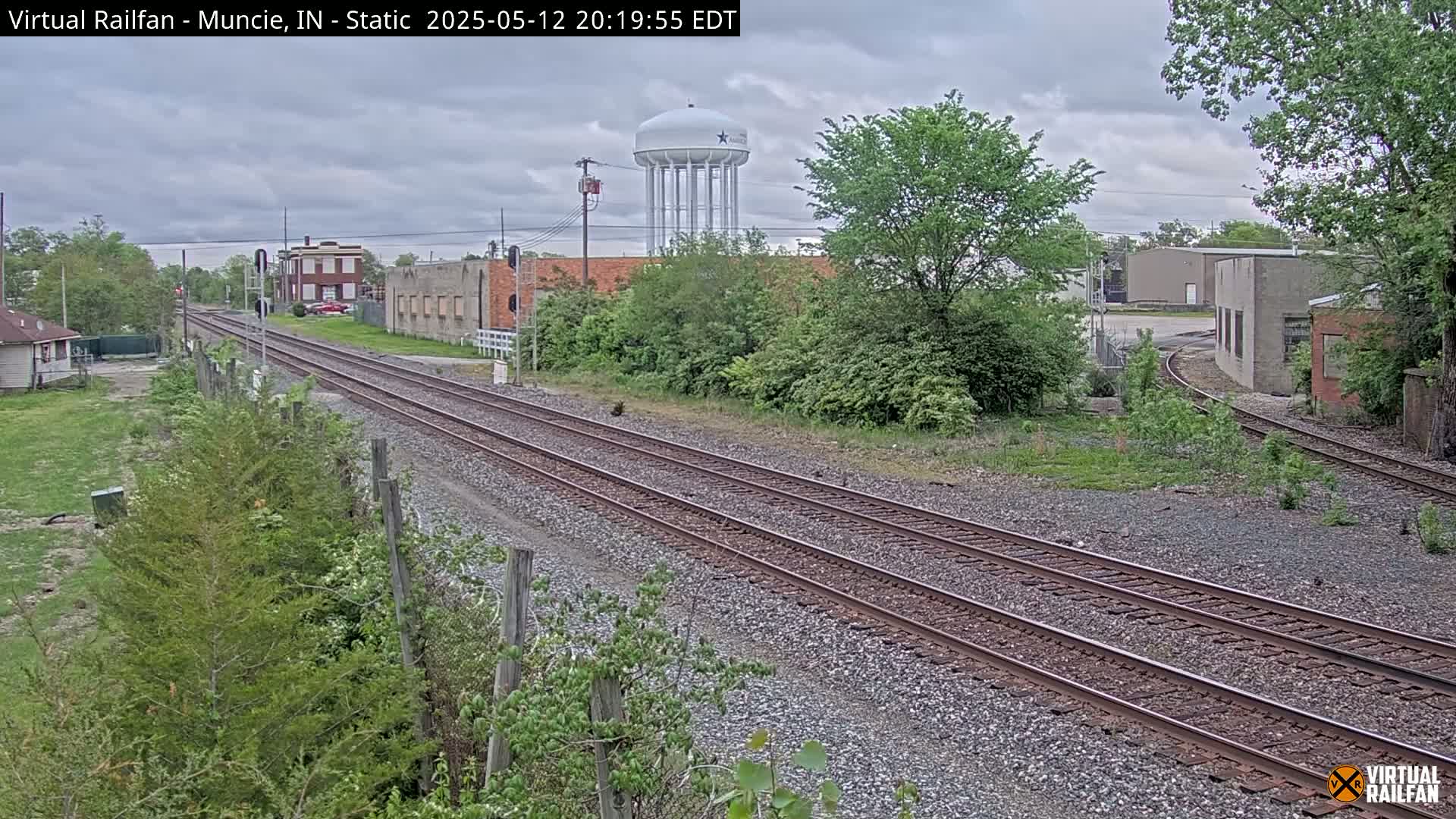 Two sets of train tracks extend into the distance under an overcast sky, passing industrial buildings and a water tower, surrounded by greenery.