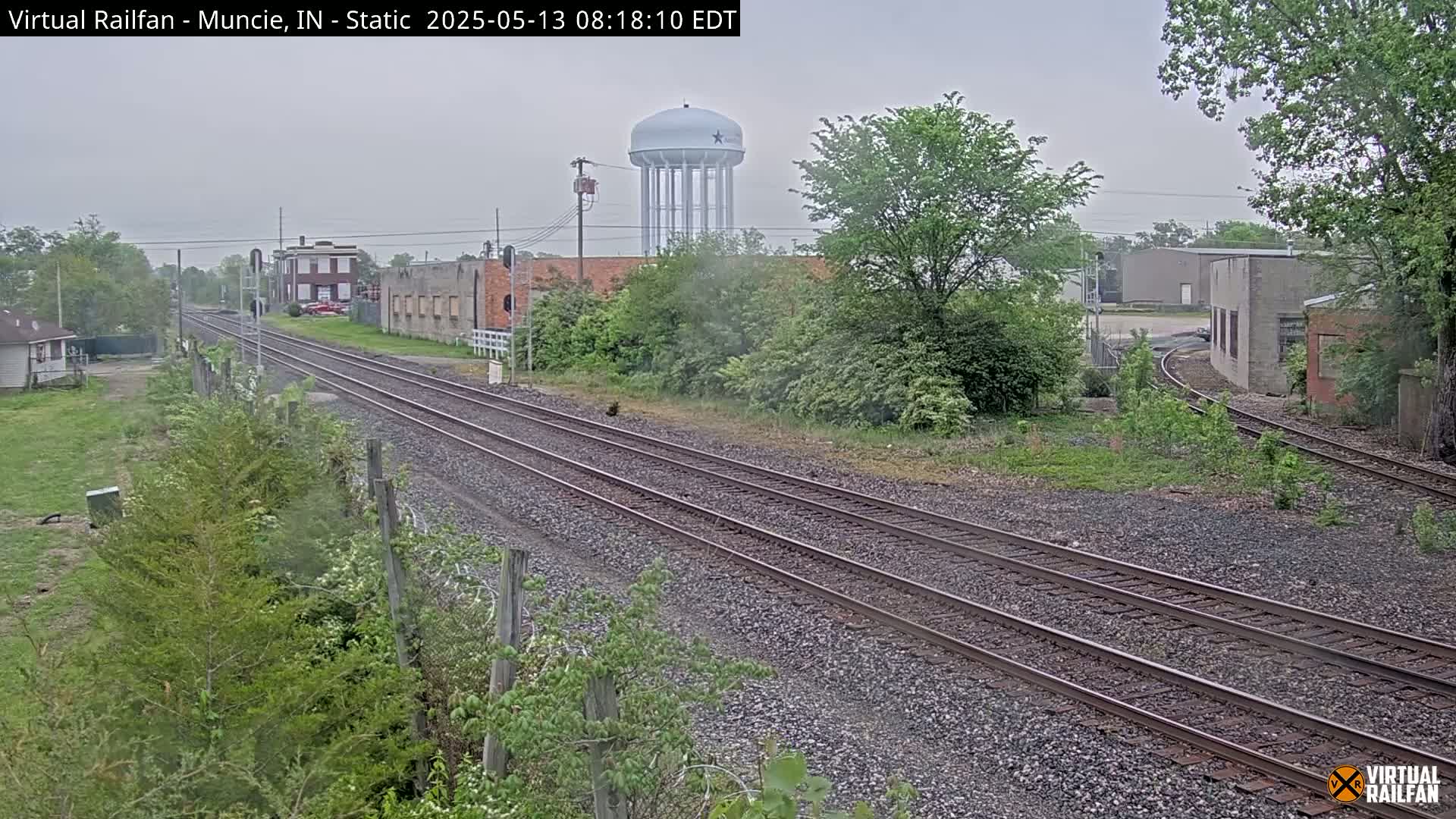 Three parallel sets of train tracks run through a town on an overcast day, past buildings and a water tower.