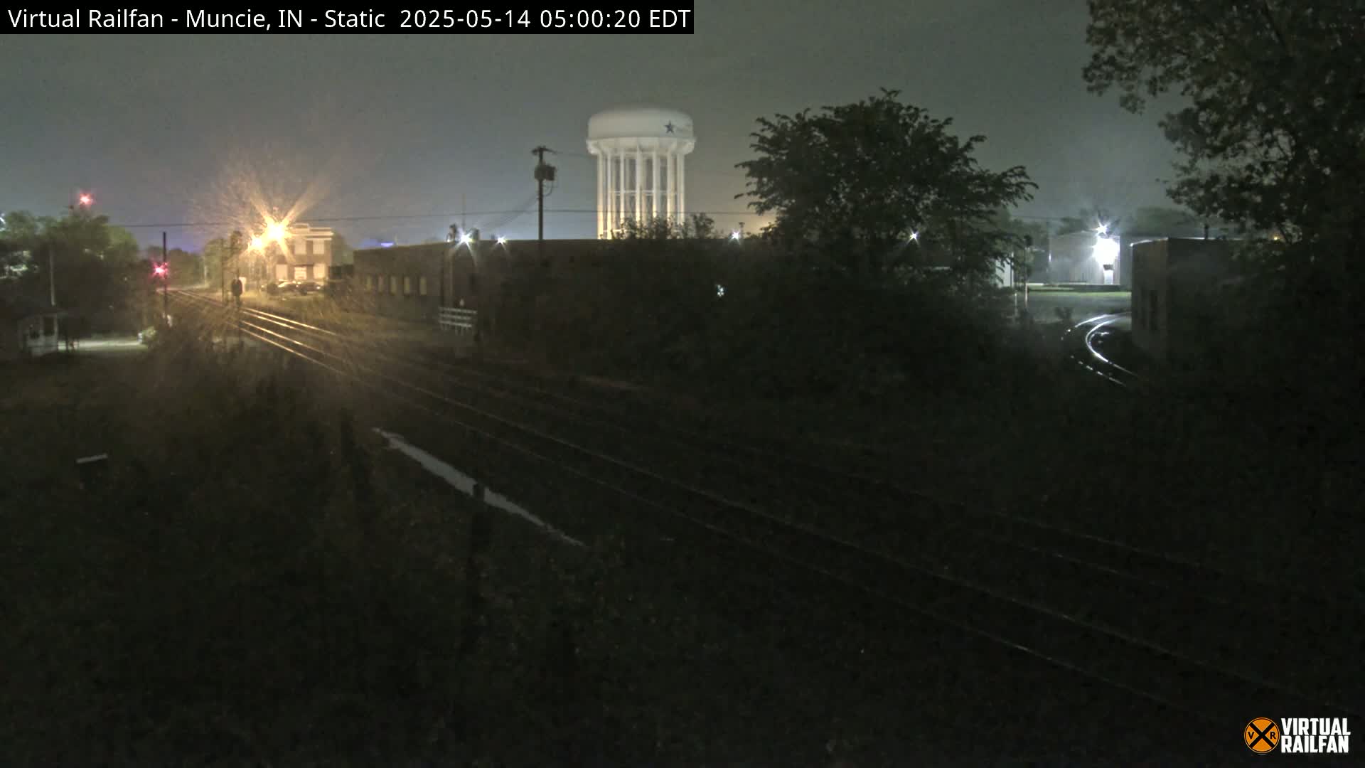 A nighttime view of rain falling on railroad tracks near a water tower and buildings.