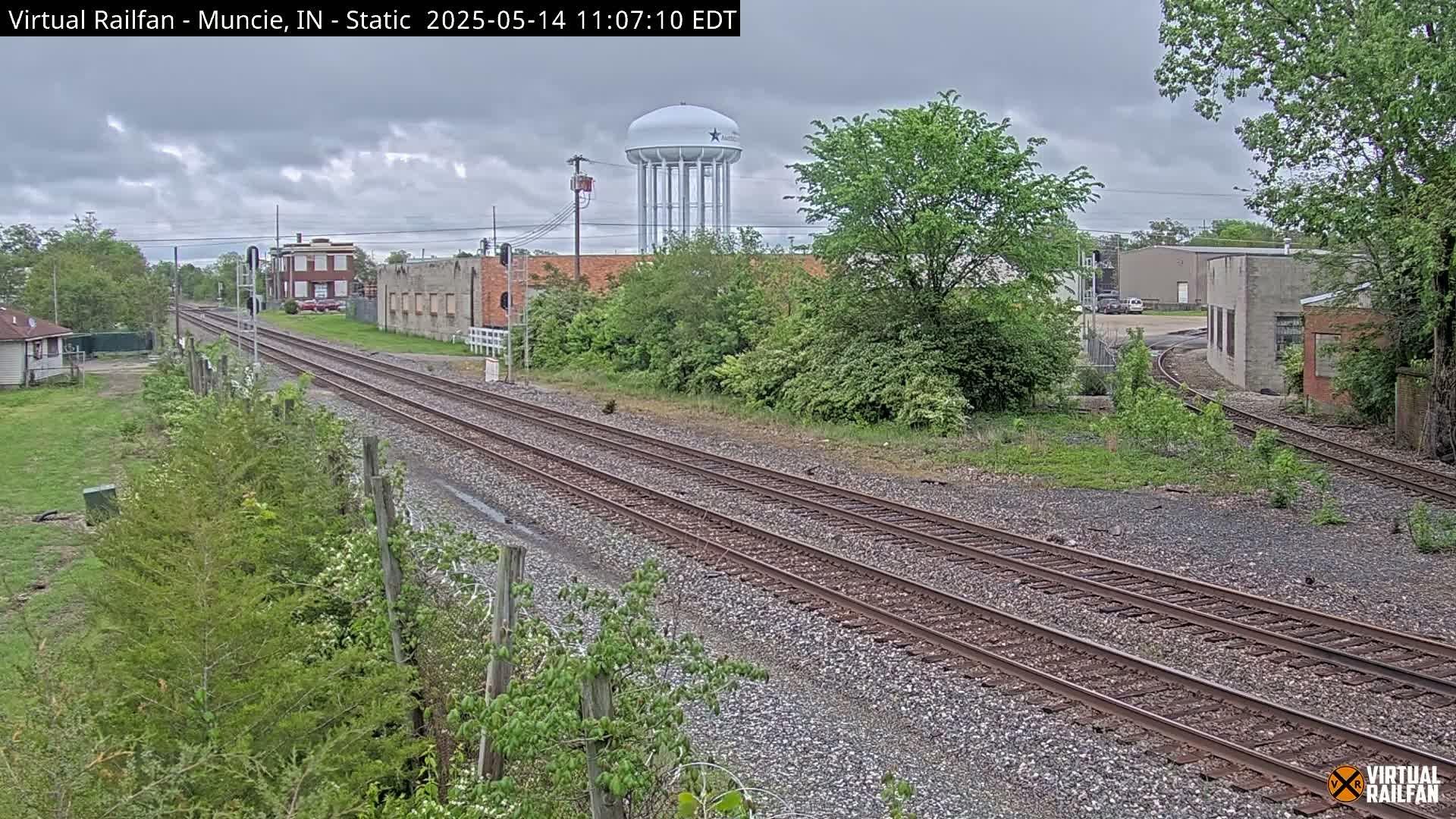 Overcast skies cover a view of railroad tracks that diverge in the distance, passing industrial buildings and a water tower, with green vegetation bordering the tracks and buildings.