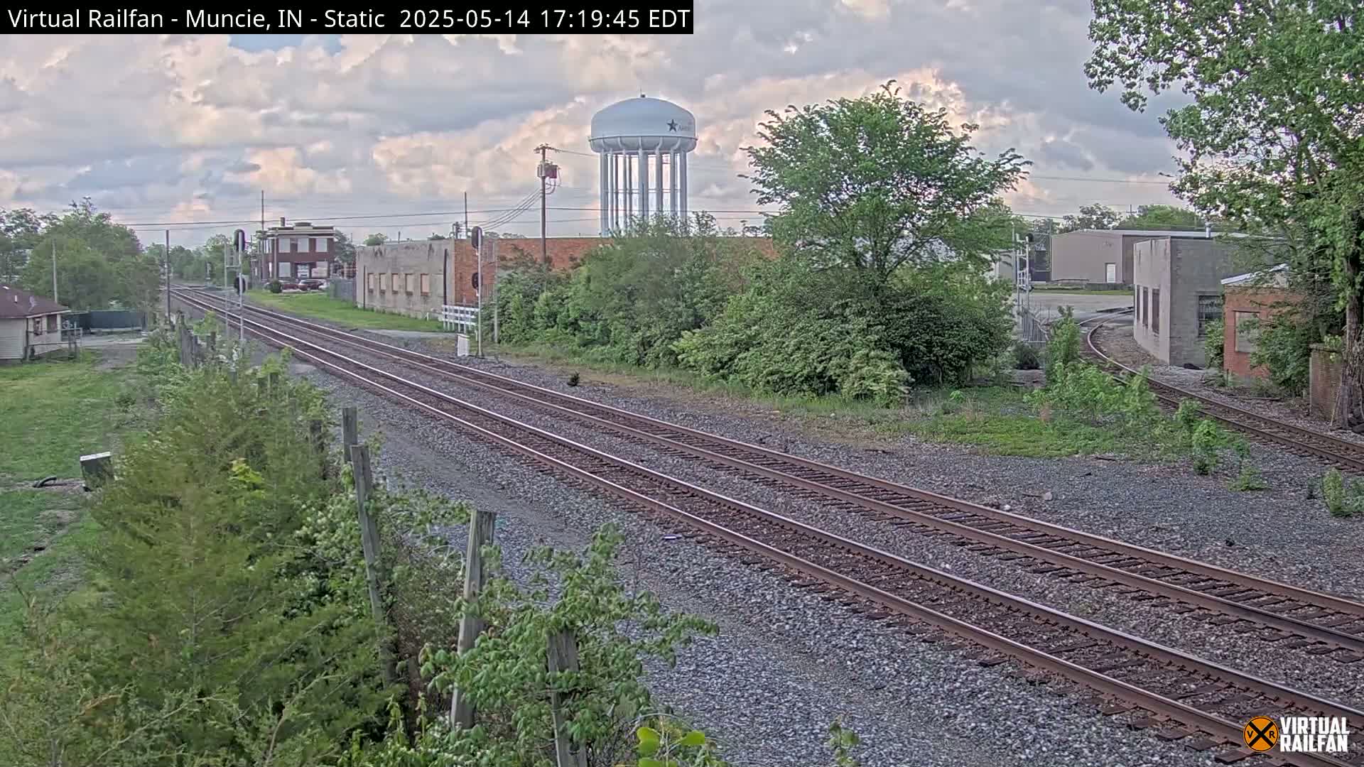 Two sets of train tracks extend into the distance under a partly cloudy sky, passing by industrial buildings and a water tower, with lush green vegetation lining the sides.