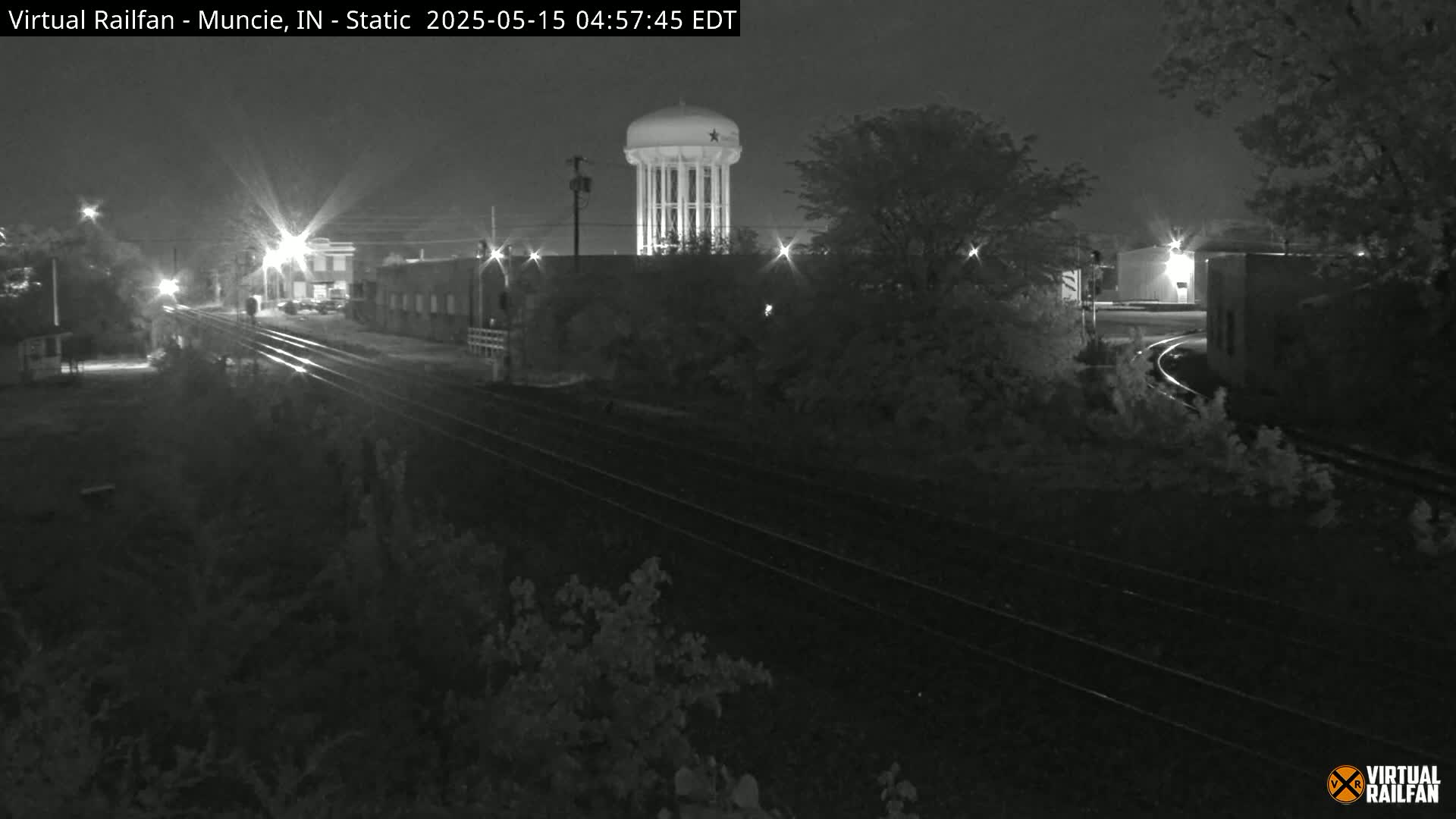 A nighttime view of railroad tracks and a water tower, with surrounding buildings and trees partially visible under dark conditions.