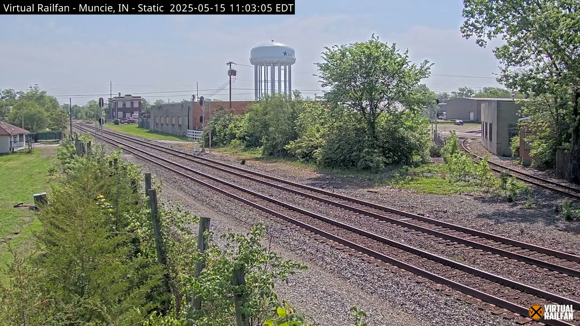 A view of railway tracks extending into the distance on a sunny day, passing industrial buildings and a water tower, surrounded by lush green vegetation.