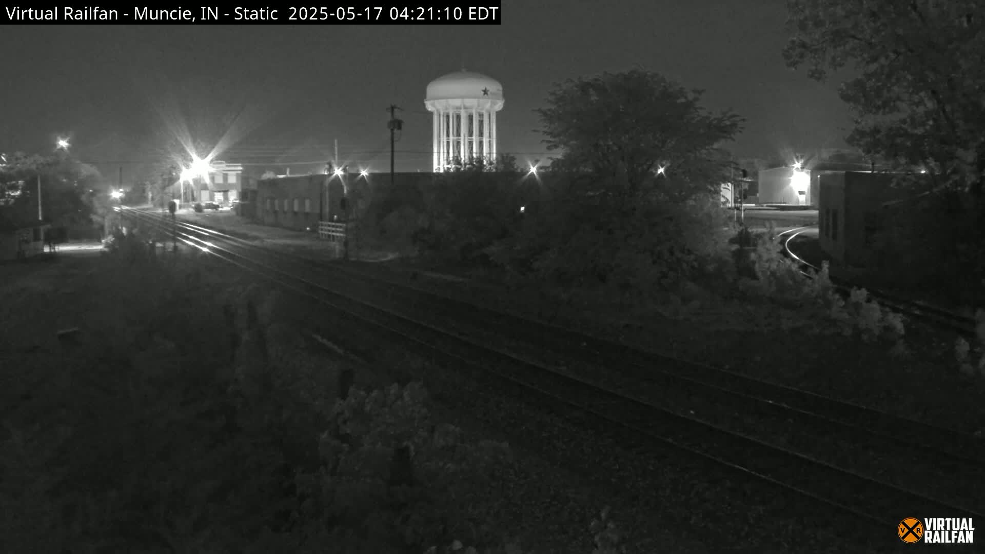 A nighttime, black-and-white view of railroad tracks leading toward a water tower and several buildings under a dark sky.