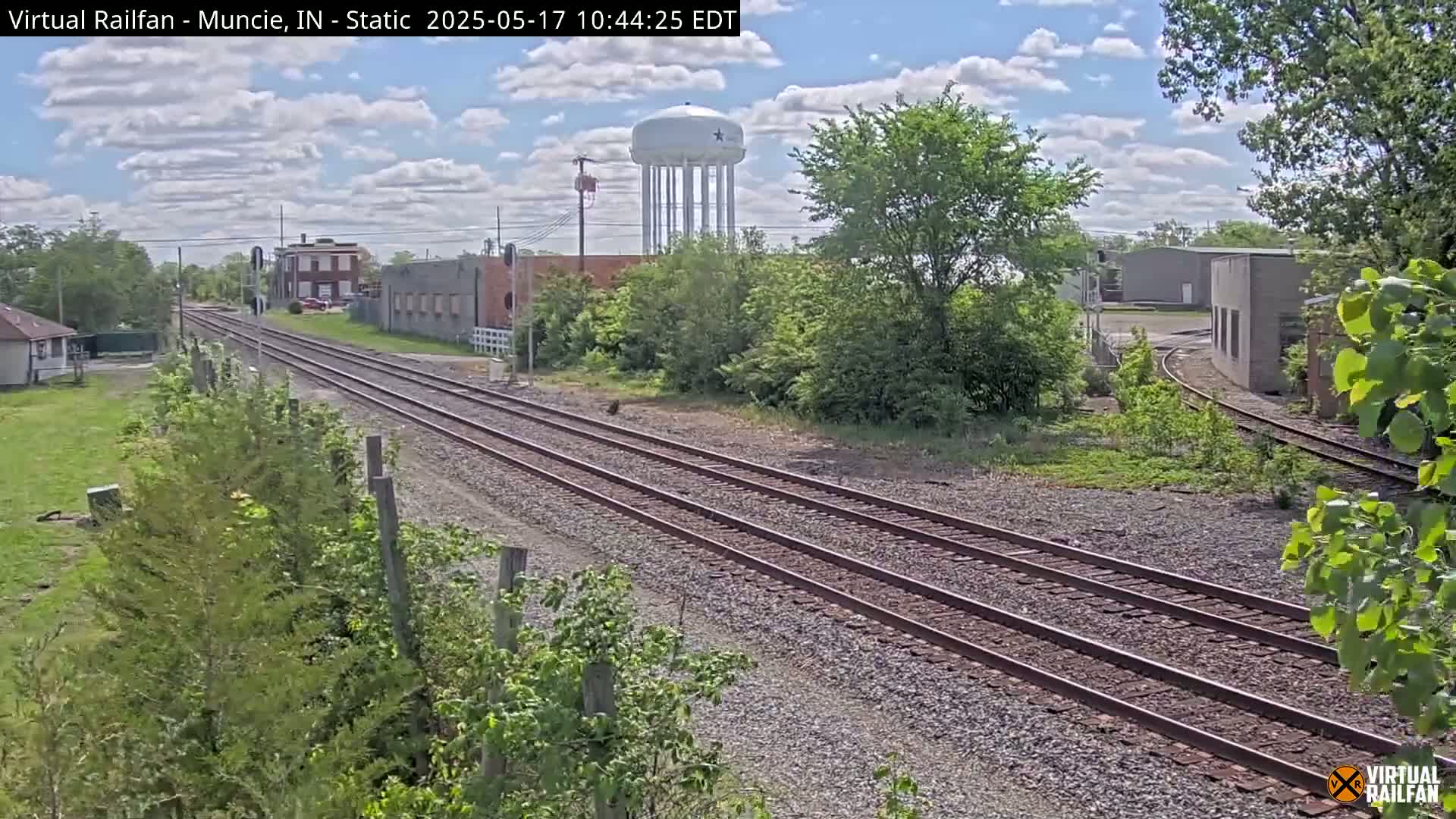 A view of railroad tracks extending into the distance under a partly cloudy sky, with buildings and a water tower visible in the background.