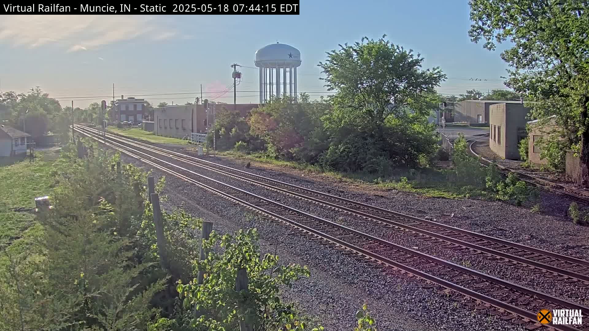 Triple train tracks extend into the distance under a clear, bright sky, passing beside a water tower and various buildings surrounded by green vegetation.