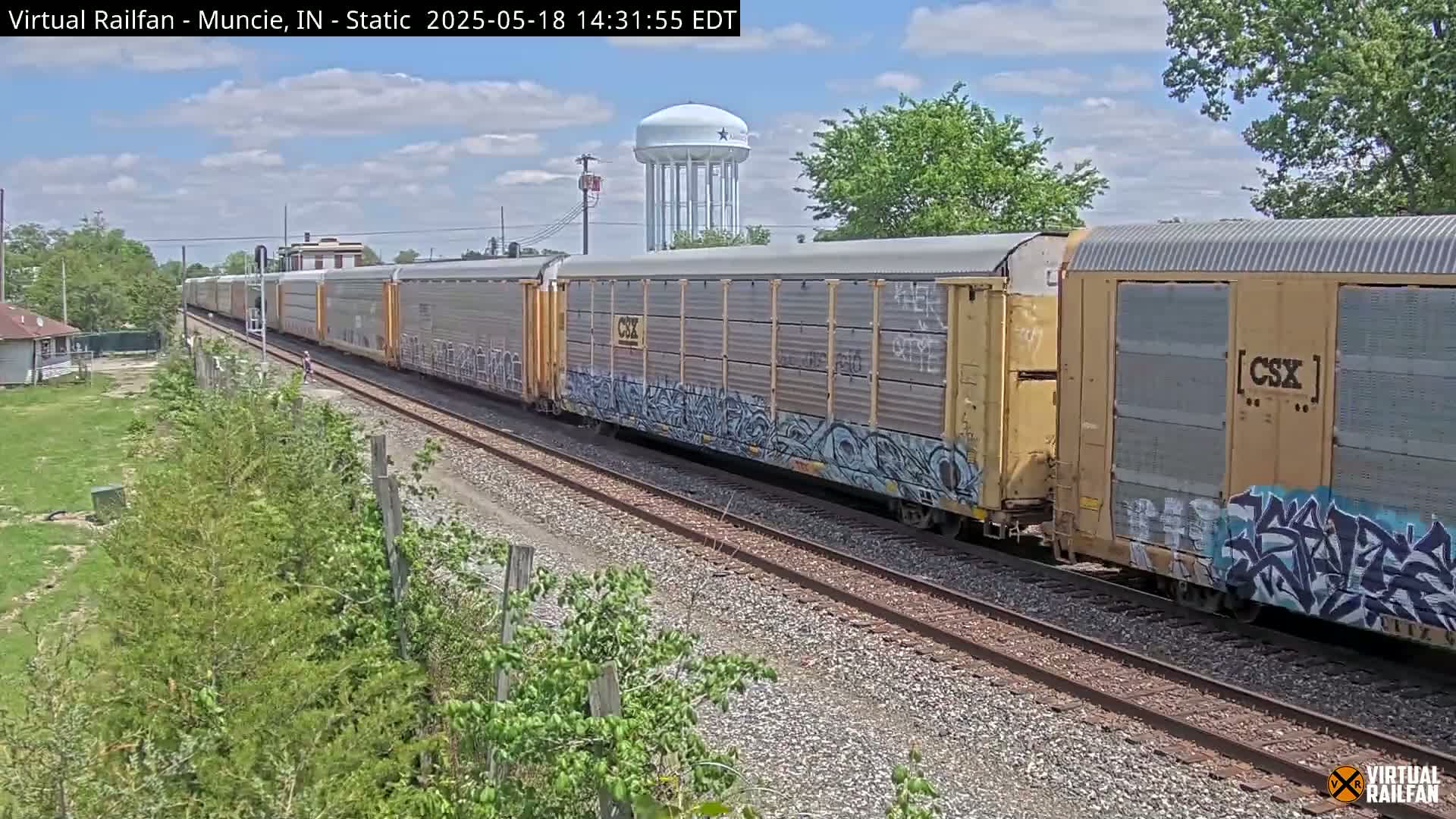 A long freight train with graffiti-covered cars passes by under partly cloudy skies.