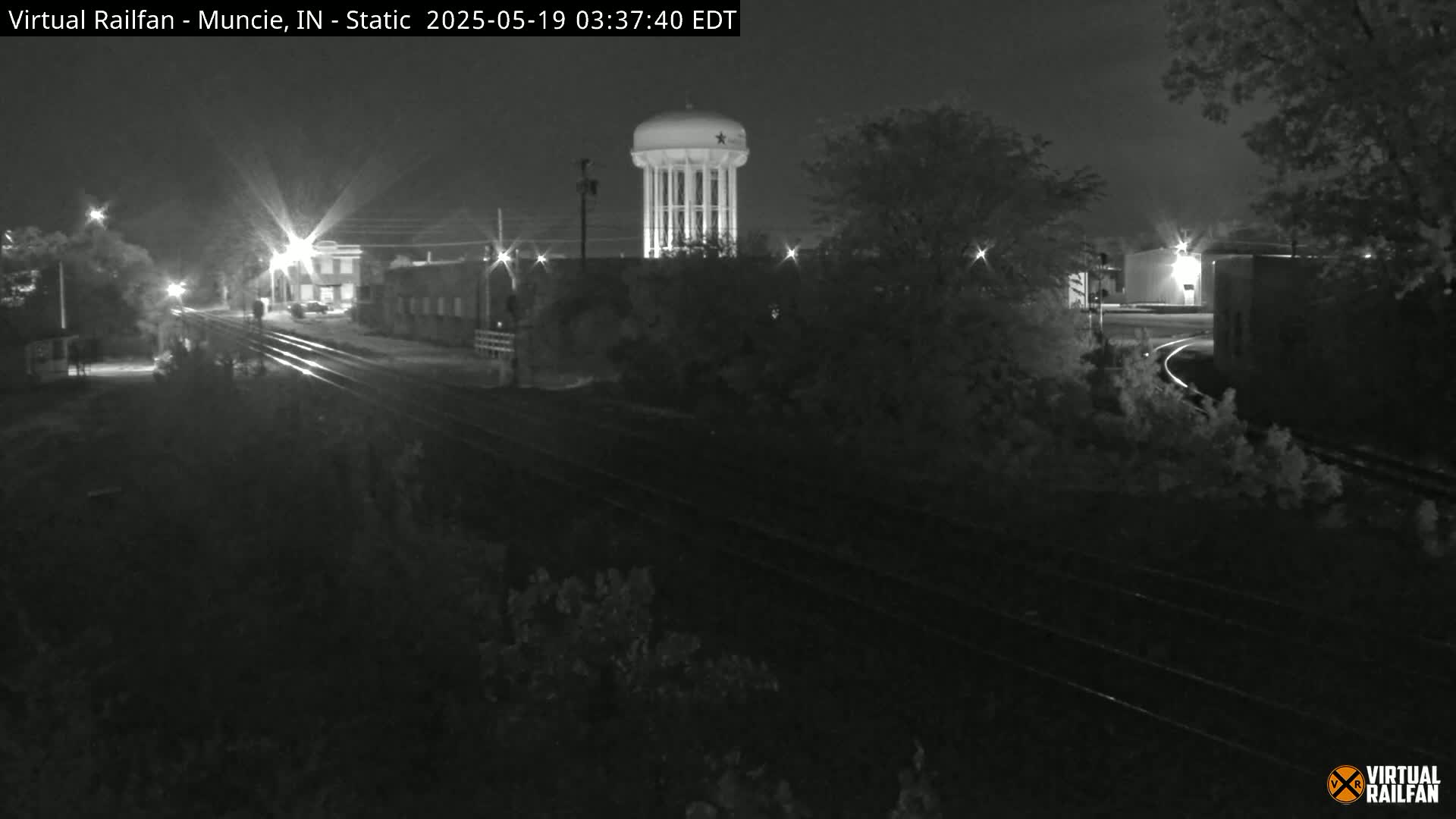 A nighttime, black-and-white view of railroad tracks leading toward a water tower and buildings under a dark sky.