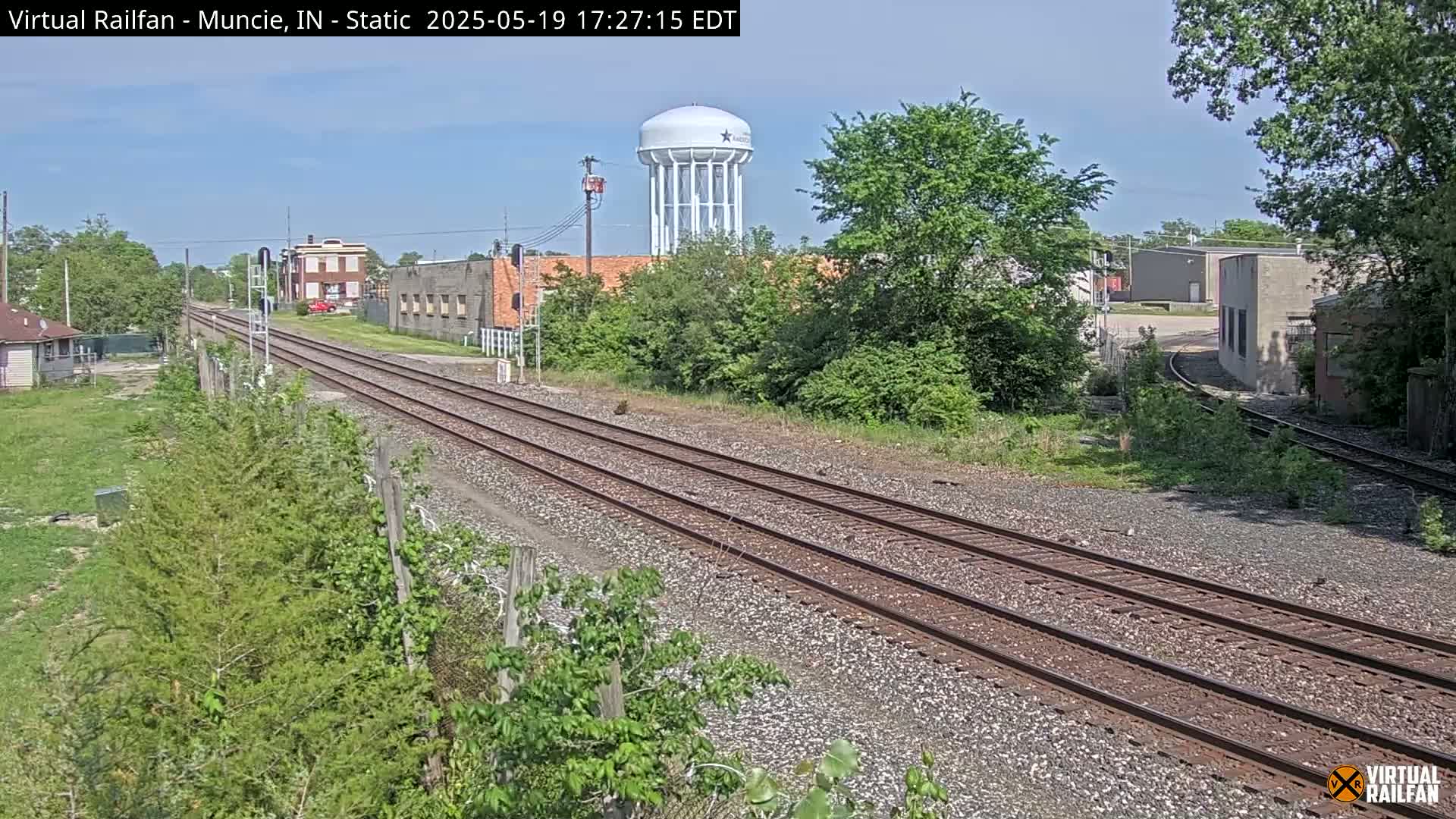 A view of train tracks extending into the distance on a sunny day, passing by buildings and a water tower amidst green vegetation.