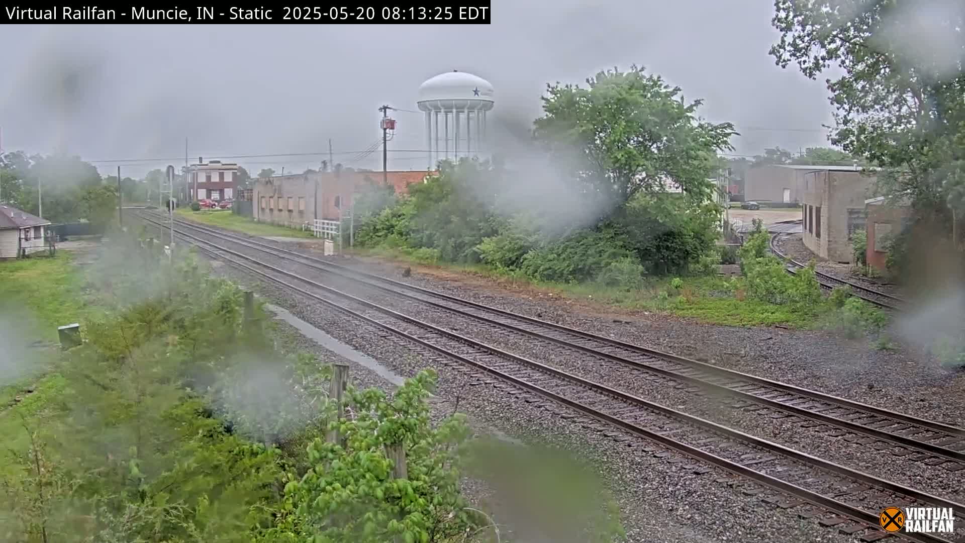 A view of railroad tracks on a rainy day, with buildings and a water tower visible in the distance.