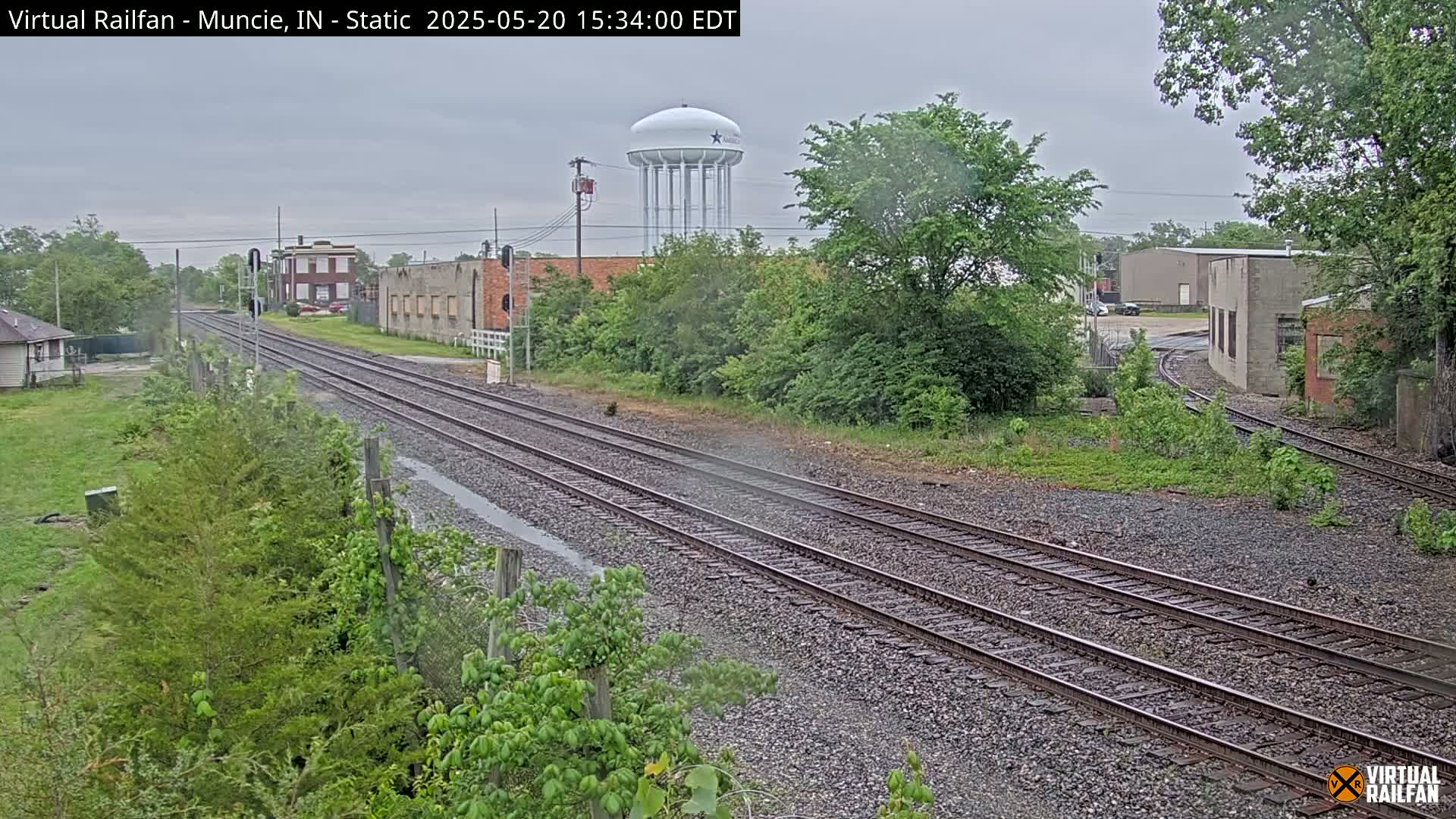 A view of railroad tracks on an overcast day, with a water tower visible in the background, surrounded by industrial buildings and vegetation.