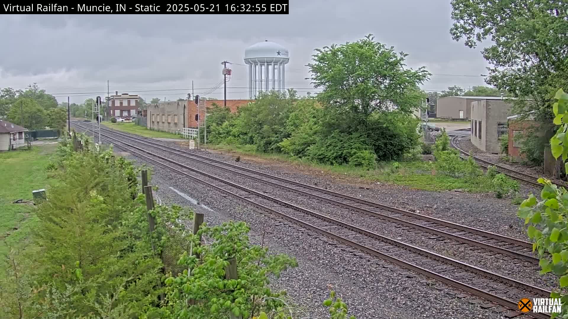 Multiple train tracks extend into the distance under an overcast sky, past industrial buildings and a water tower, surrounded by lush green vegetation.