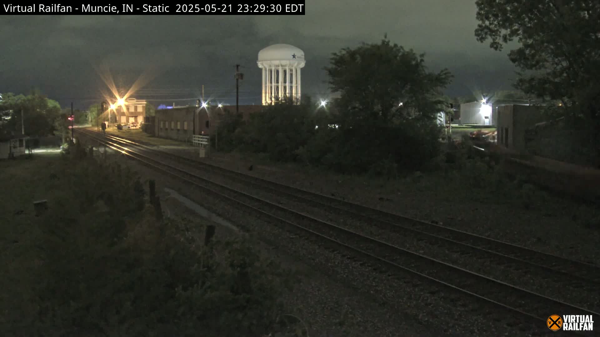 A nighttime view of train tracks, alongside buildings and a water tower, under a dark, cloudy sky.