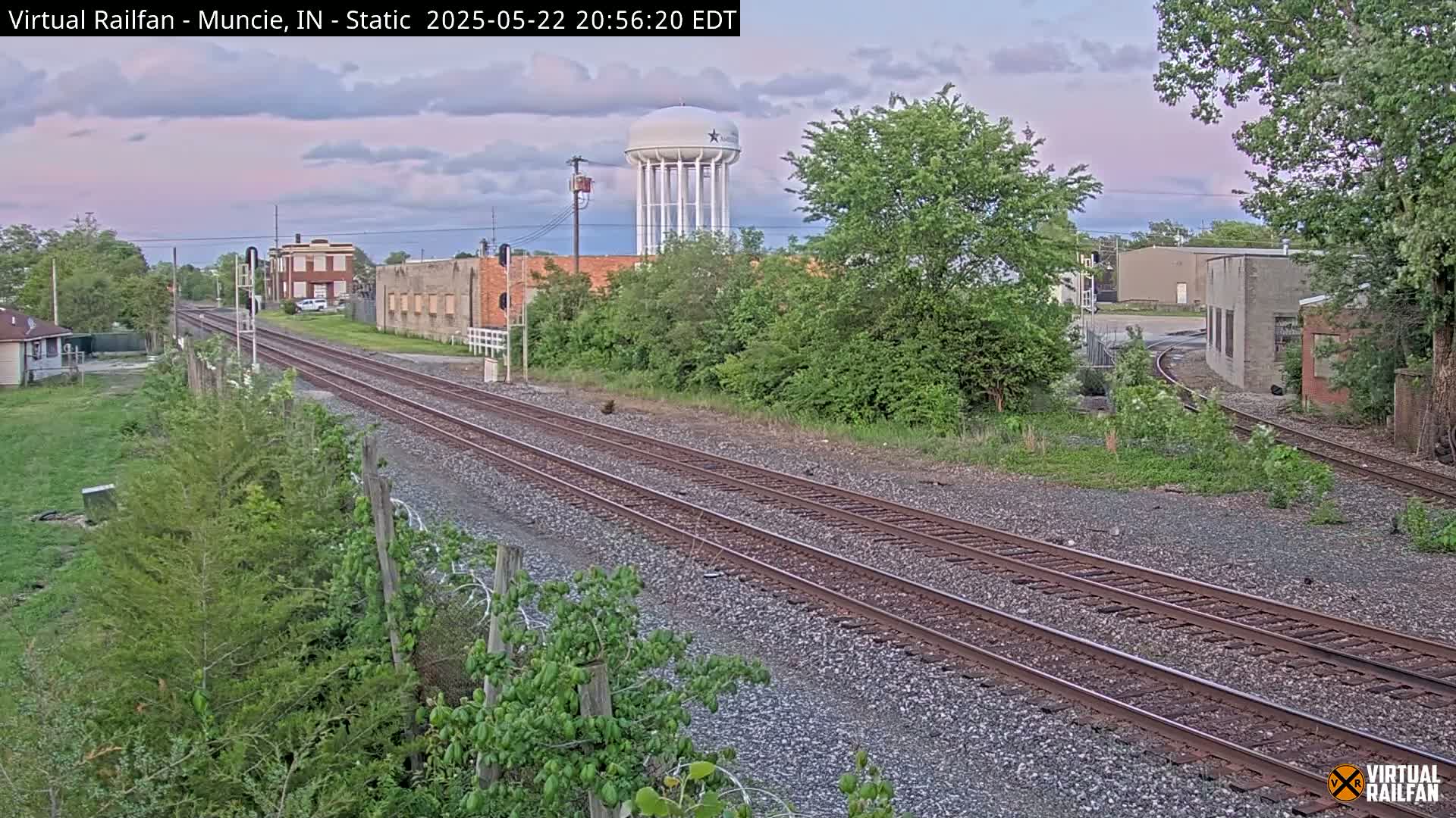 Two sets of train tracks extend into the distance under a partly cloudy evening sky, past several buildings and a water tower.