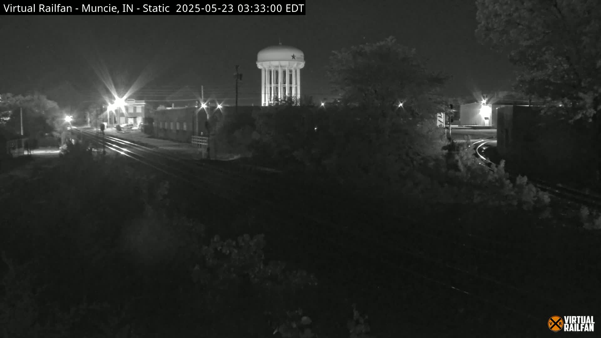 A nighttime, black-and-white view of railroad tracks leading towards a water tower, with buildings and trees visible in the background.
