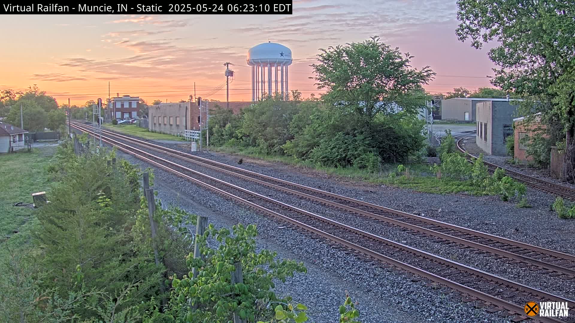 Railroad tracks extend into the distance past industrial buildings and a water tower under a sunrise sky.