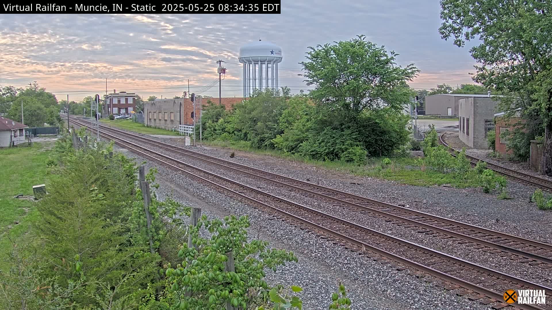 Two sets of train tracks extend into the distance past industrial buildings and a water tower under a partly cloudy sky.