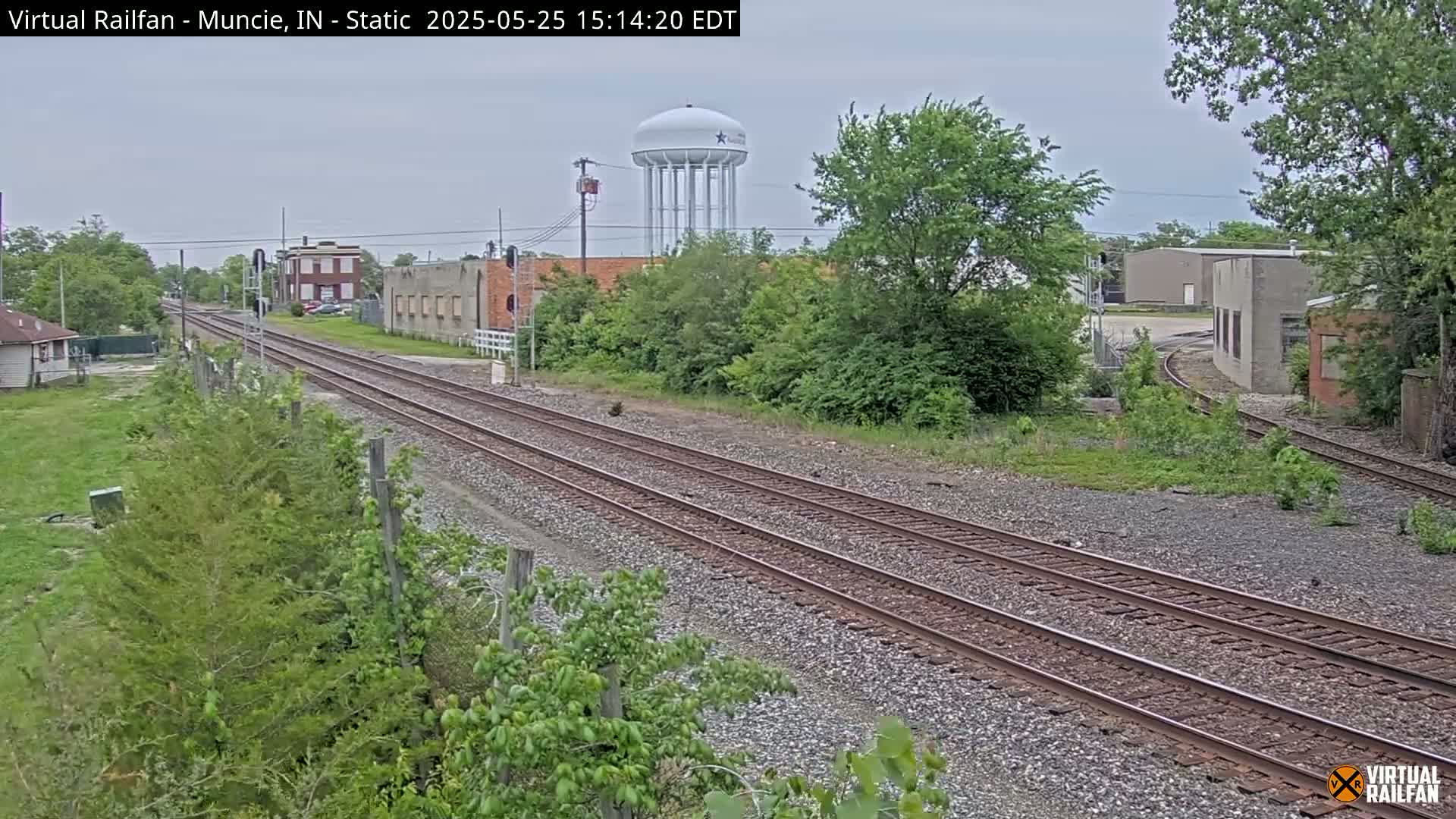 A view of railroad tracks extending into the distance under an overcast sky, passing alongside industrial buildings and a water tower, with lush green vegetation bordering the tracks.