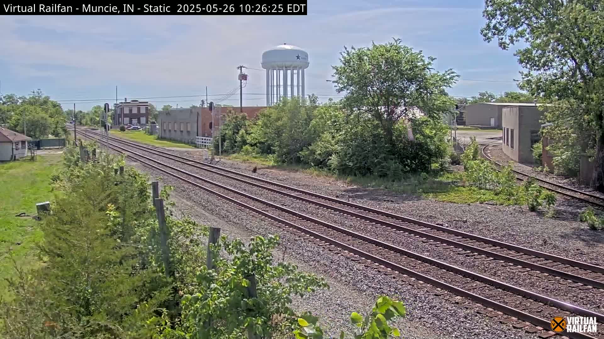 Under a mostly sunny sky, multiple train tracks extend into the distance past industrial buildings and a water tower.