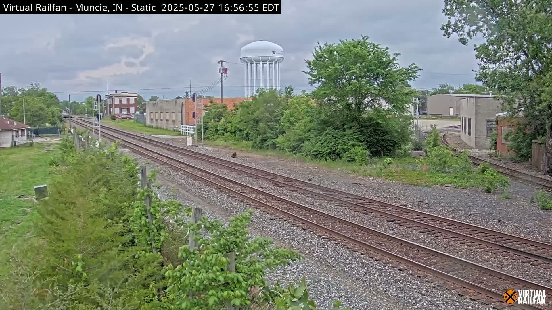 Overcast skies loom over a railroad track that winds through a town, passing industrial buildings and a water tower.