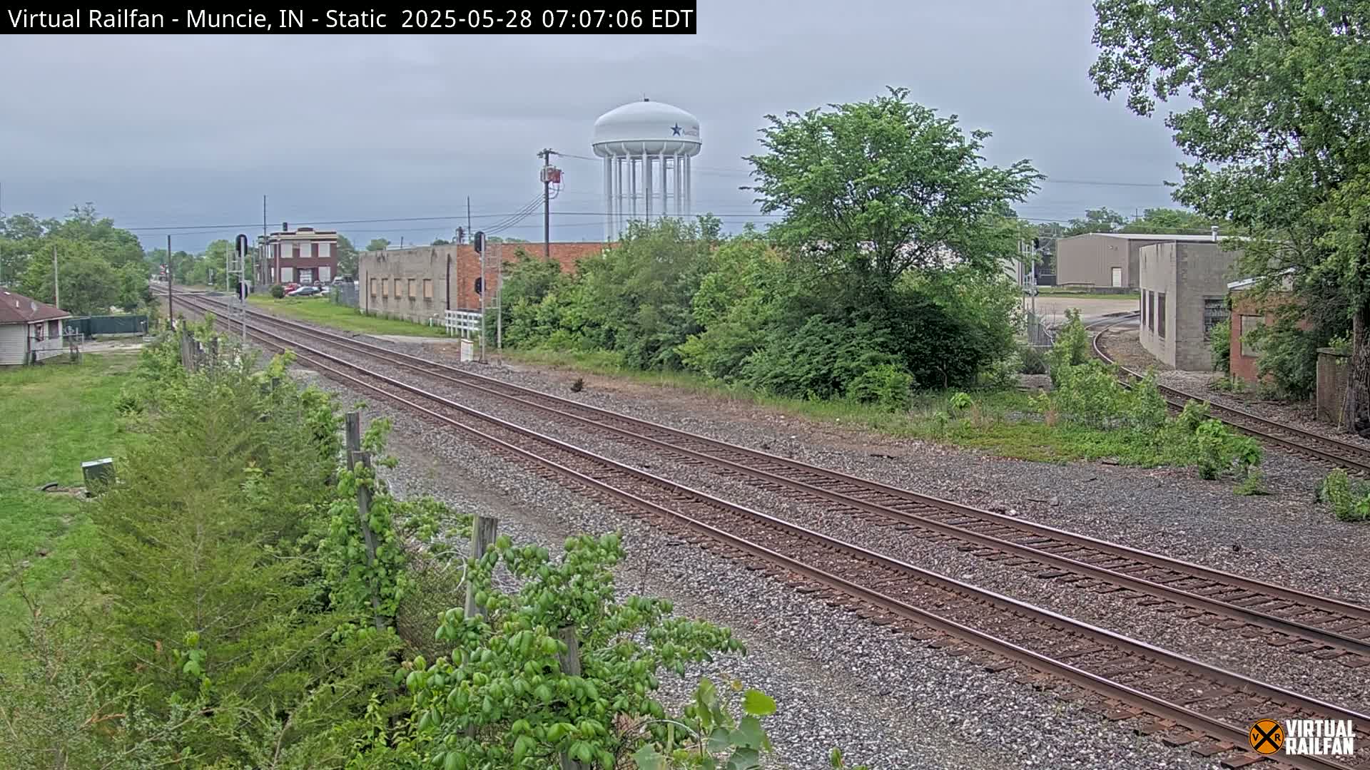 A view of railroad tracks on an overcast day, with industrial buildings, a water tower, and lush green vegetation visible in the background.