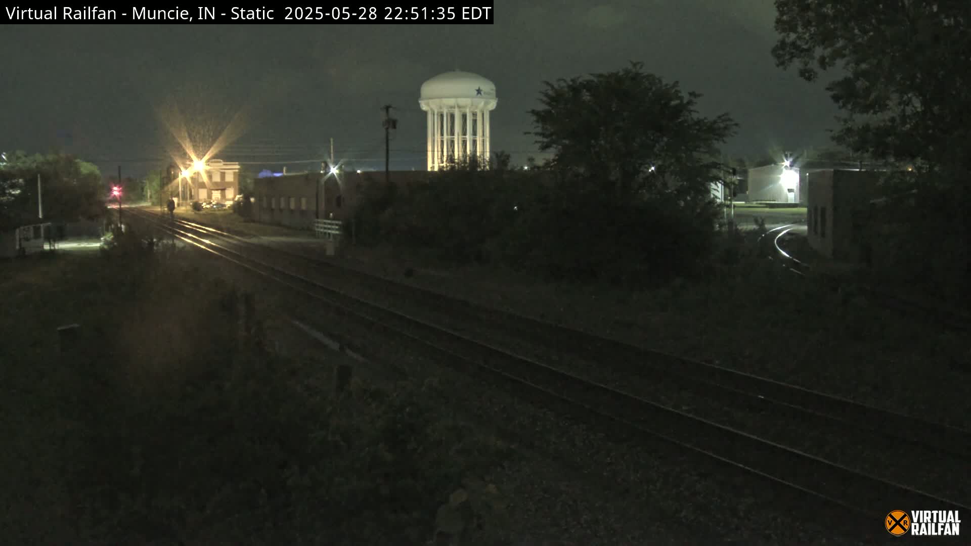 At night, railroad tracks stretch into the distance, passing buildings and a water tower under a dark sky.