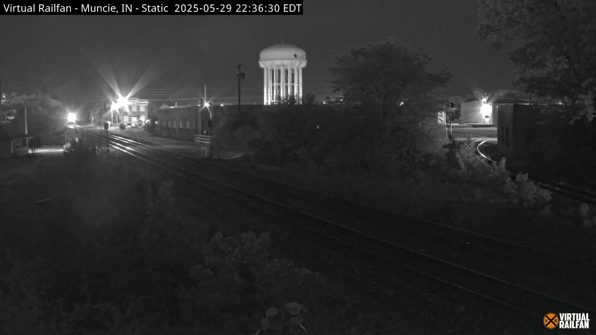 A nighttime, black-and-white view of railroad tracks and buildings, including a water tower, under a clear sky.