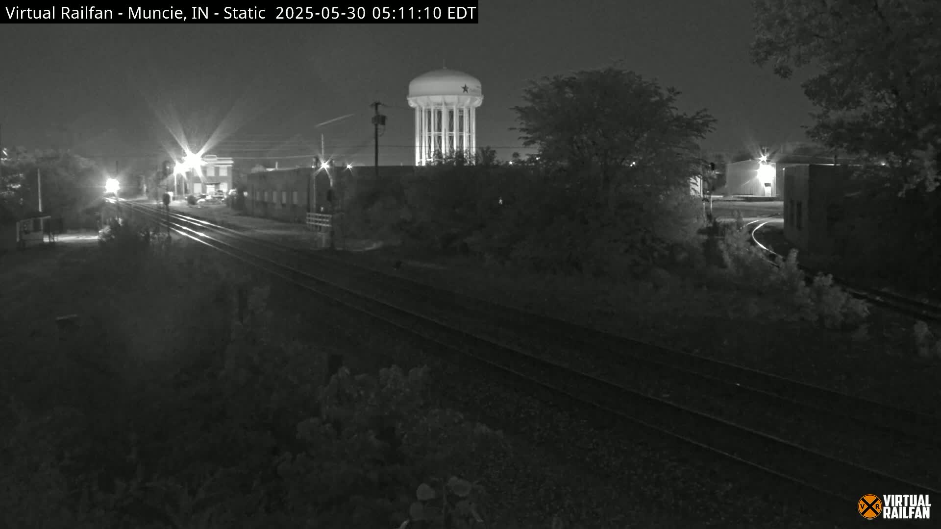A nighttime black and white view of railroad tracks, buildings, and a water tower under a clear sky.