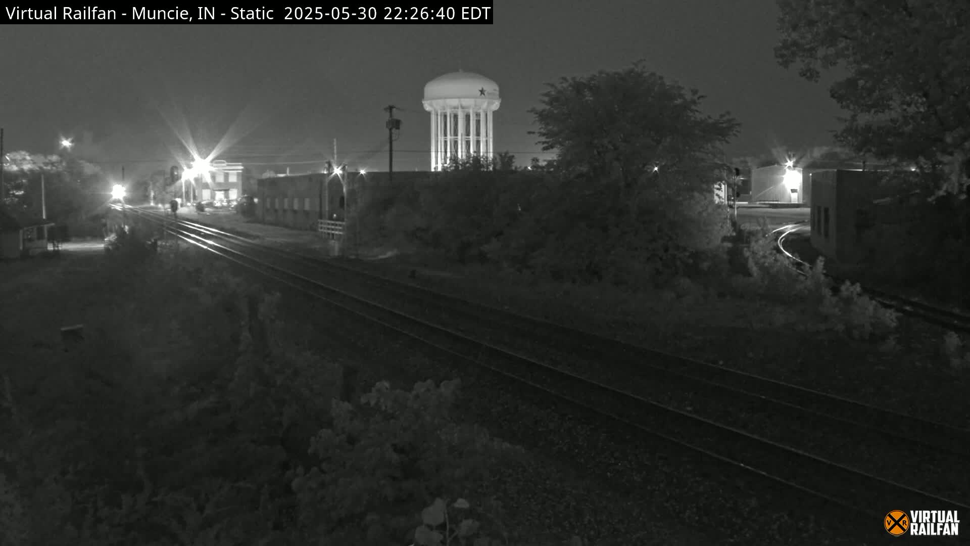 A nighttime, black and white view of train tracks that curve to the right, passing by buildings and a water tower under a dark sky.