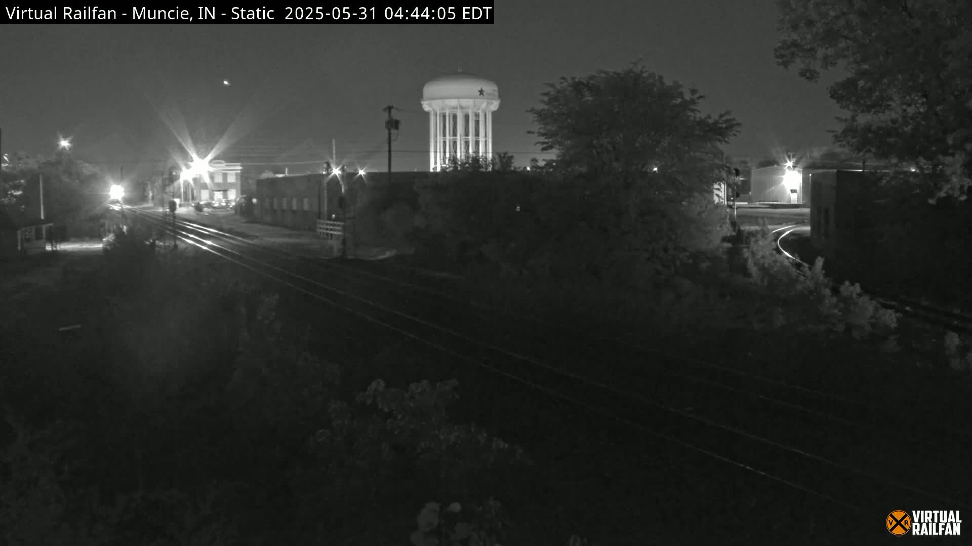 A nighttime, black and white view of railroad tracks, buildings, and a water tower under a clear sky.
