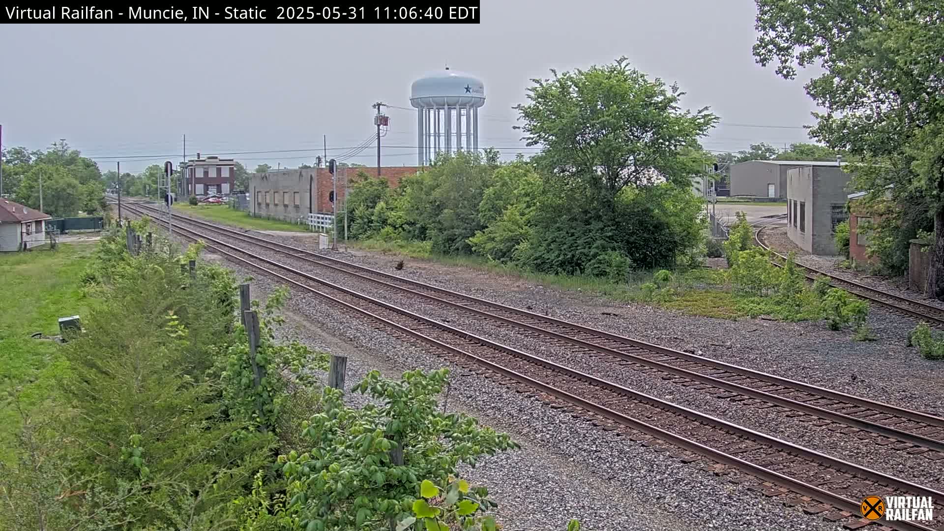 Under an overcast sky, a view of railroad tracks extends into the distance, passing by industrial buildings and a water tower, with lush green vegetation lining the sides.