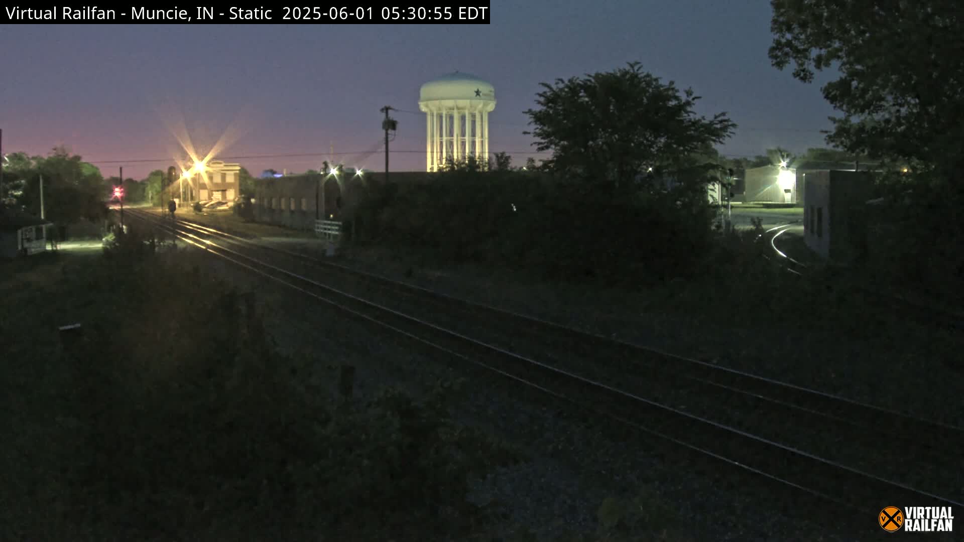 Under a twilight sky, railroad tracks extend into the distance past dimly lit buildings, towards a water tower.