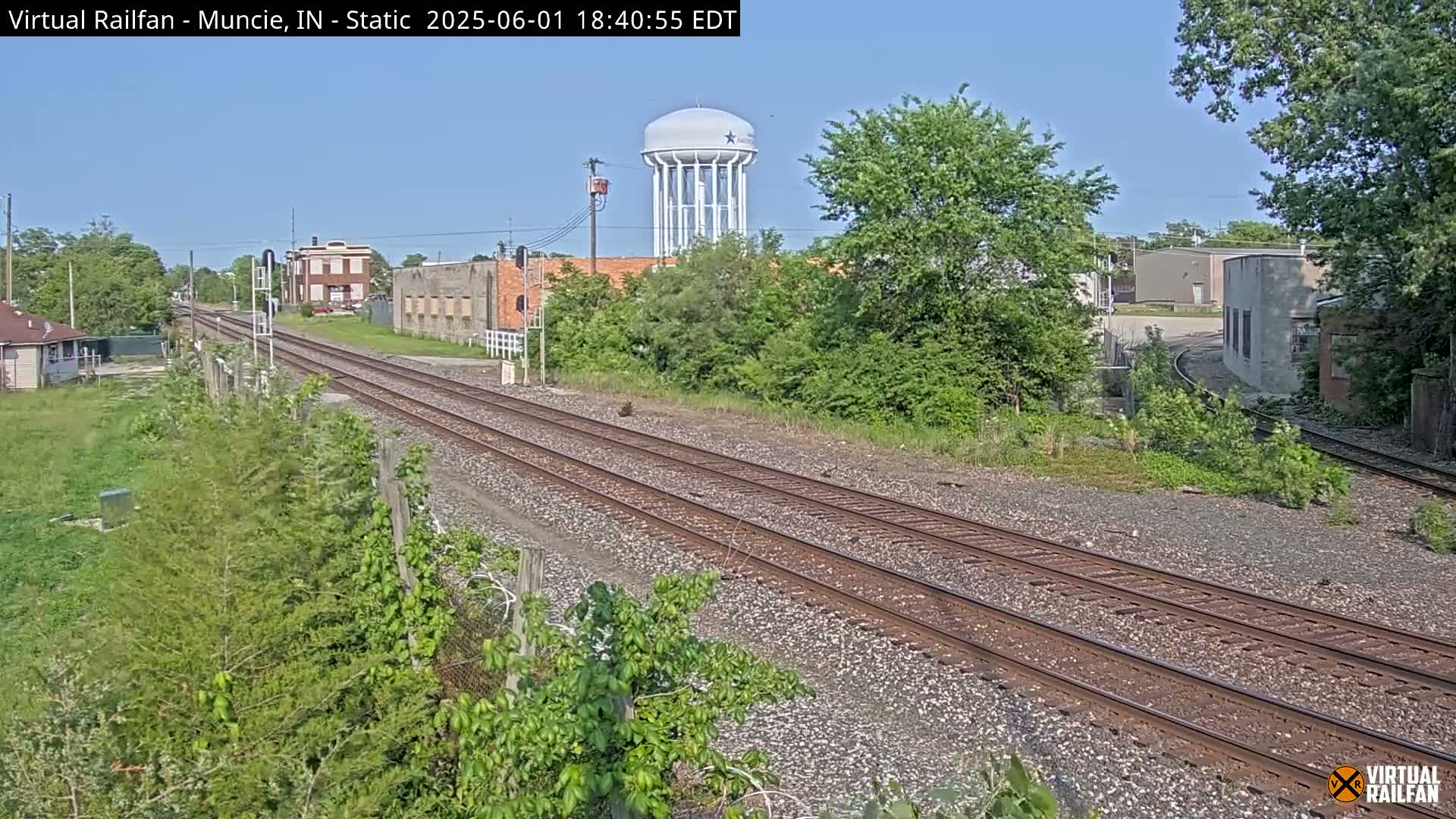 Two sets of train tracks stretch into the distance under a clear, sunny sky, past some buildings and lush green vegetation, with a water tower visible in the background.