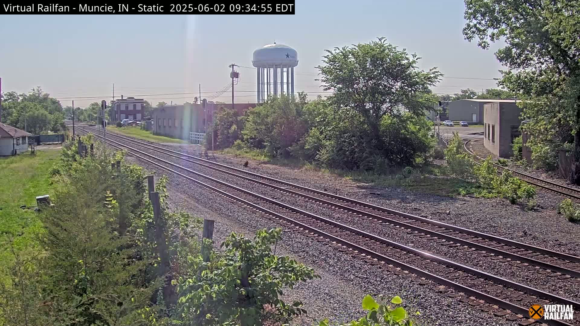 A sunny day view of multiple sets of train tracks, industrial buildings, and a water tower, surrounded by green vegetation.