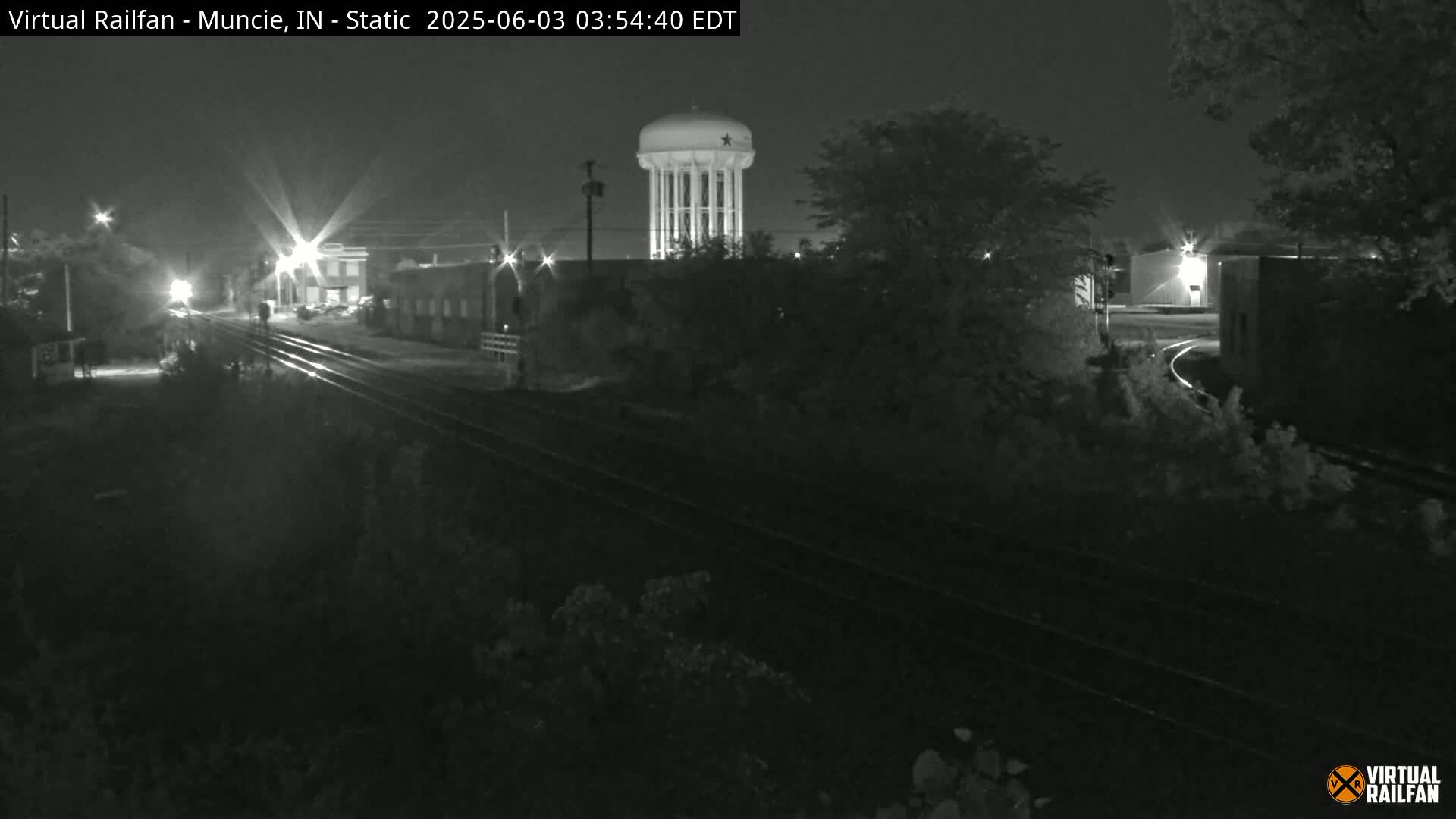 A nighttime, black-and-white view of railroad tracks intersecting with a road, featuring a water tower in the background under a clear sky.