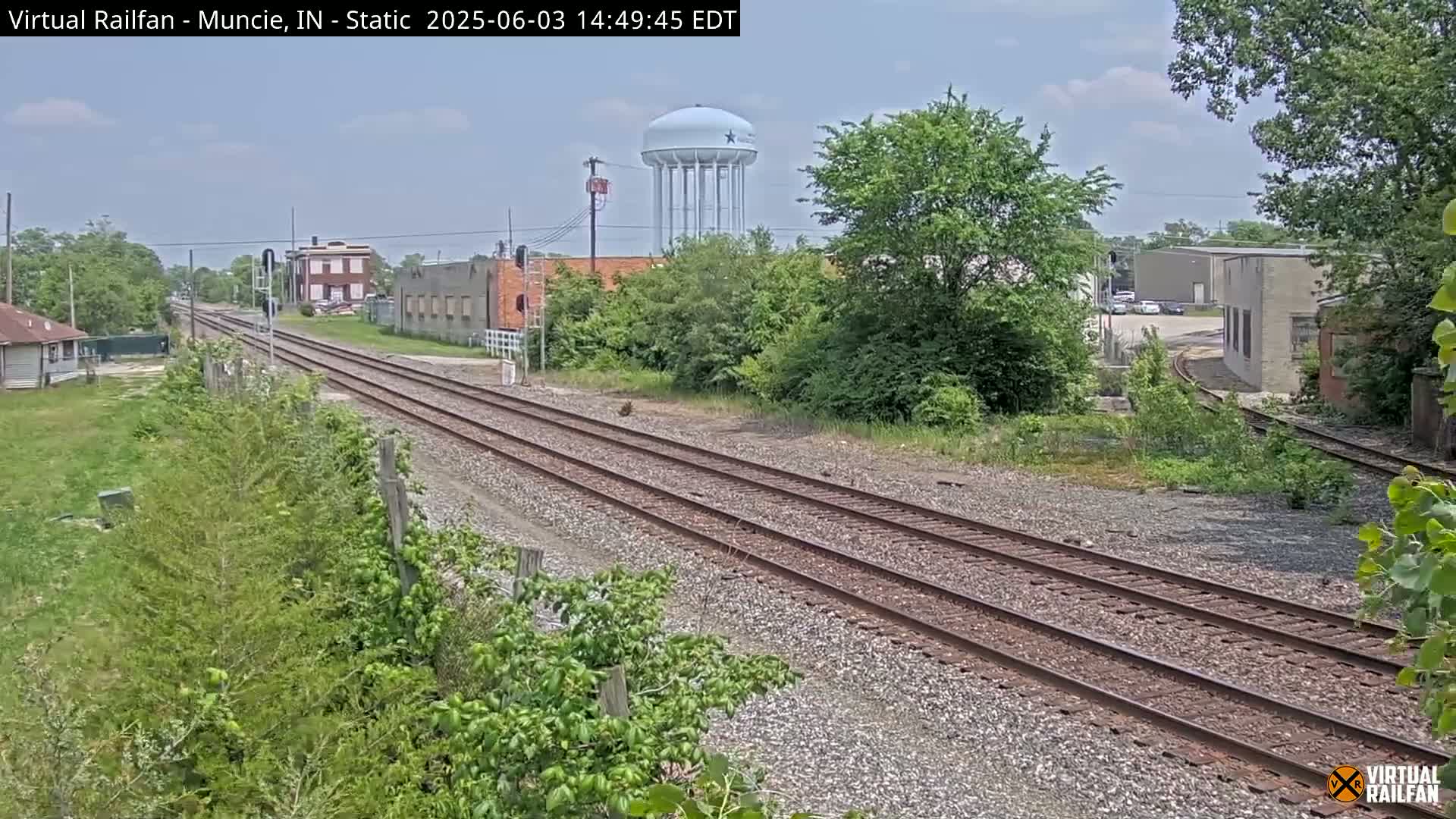 A view of railroad tracks extending into the distance under a partly cloudy sky, passing by industrial buildings and a water tower, surrounded by green vegetation.