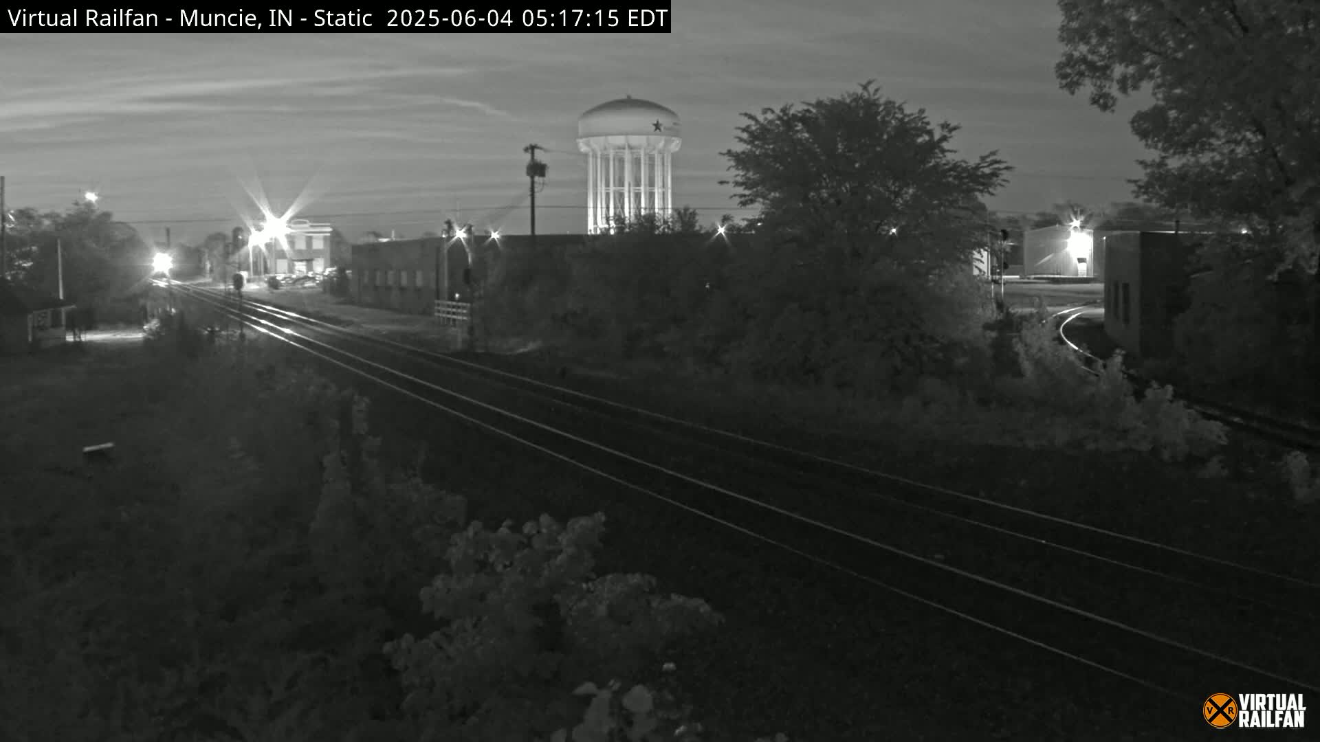 A nighttime, grayscale view of railroad tracks winding through a landscape of buildings and trees, with a water tower visible in the background under a dark sky.