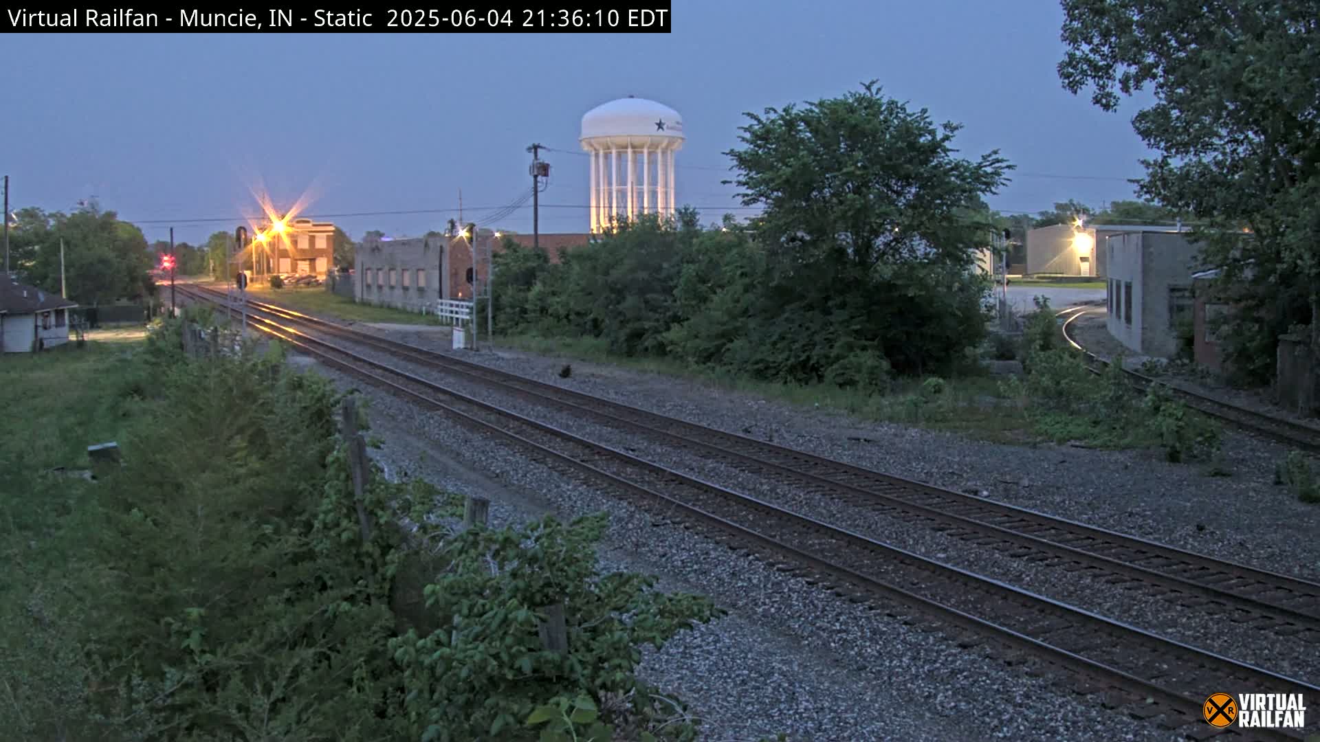 Under a twilight sky, railroad tracks stretch toward a water tower and industrial buildings, partially illuminated by streetlights.