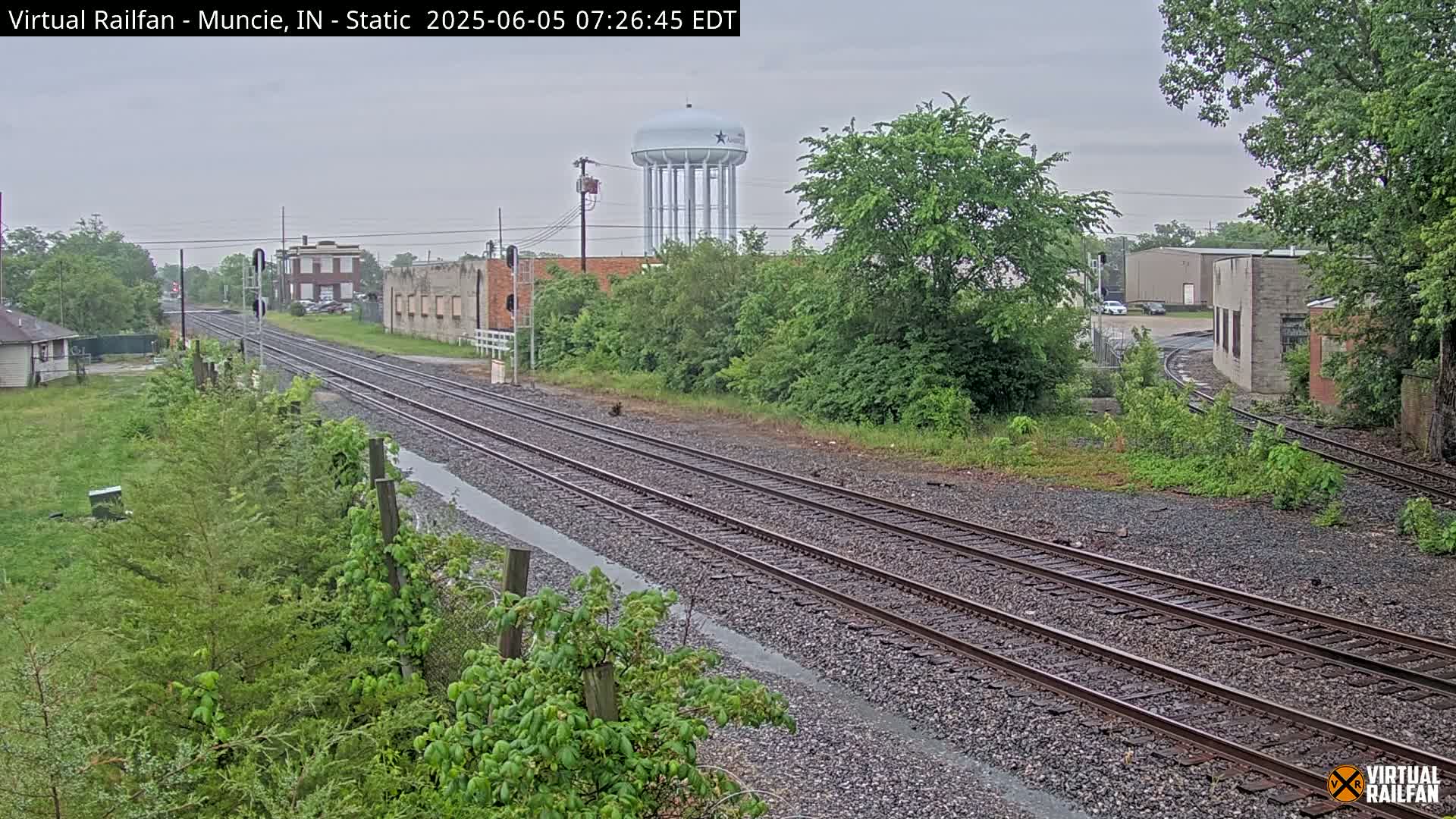 A view of railroad tracks on an overcast day, with some buildings and a water tower visible in the distance.