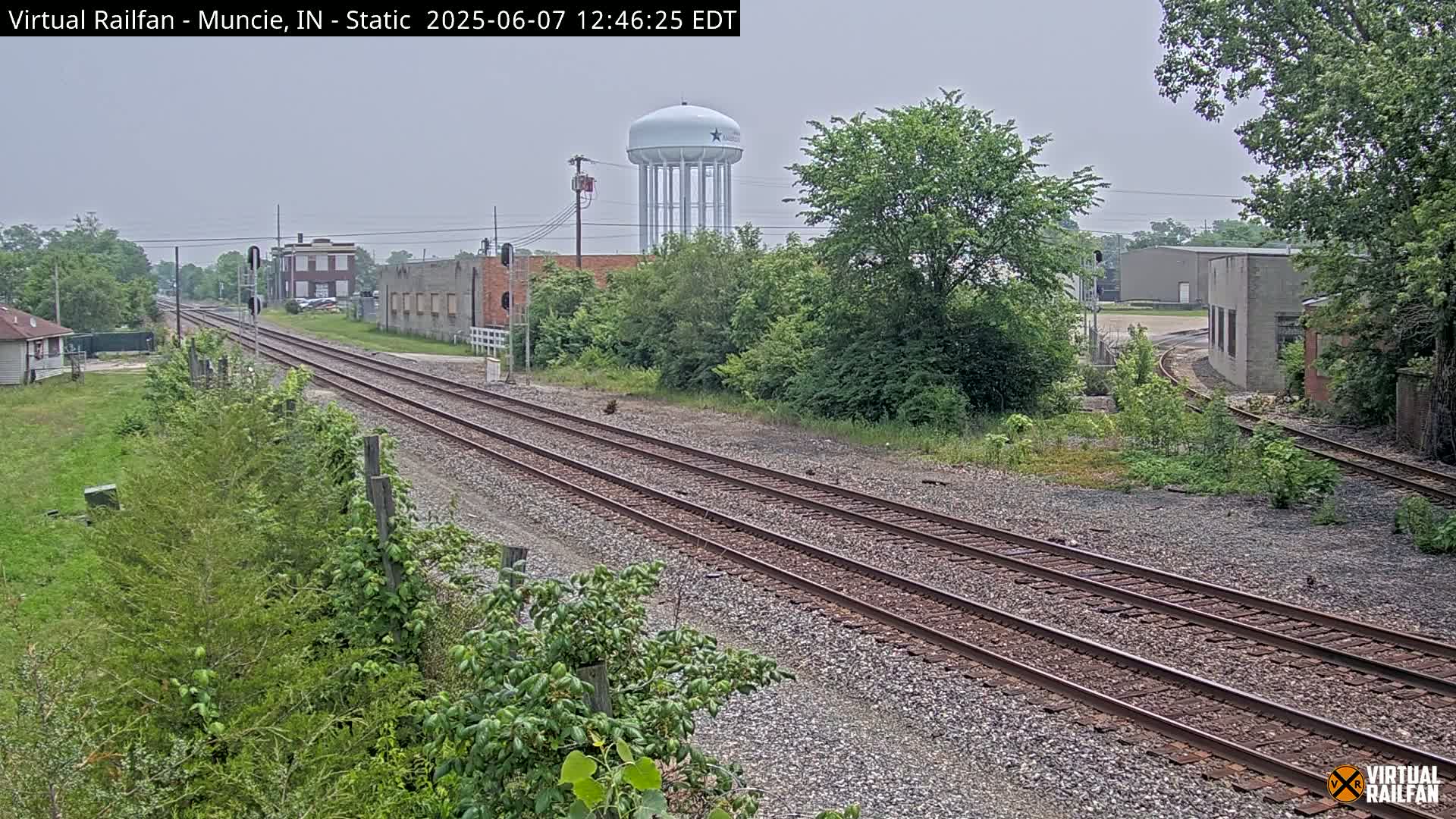 A view of railroad tracks splitting into two paths, with buildings and a water tower in the background under an overcast sky.