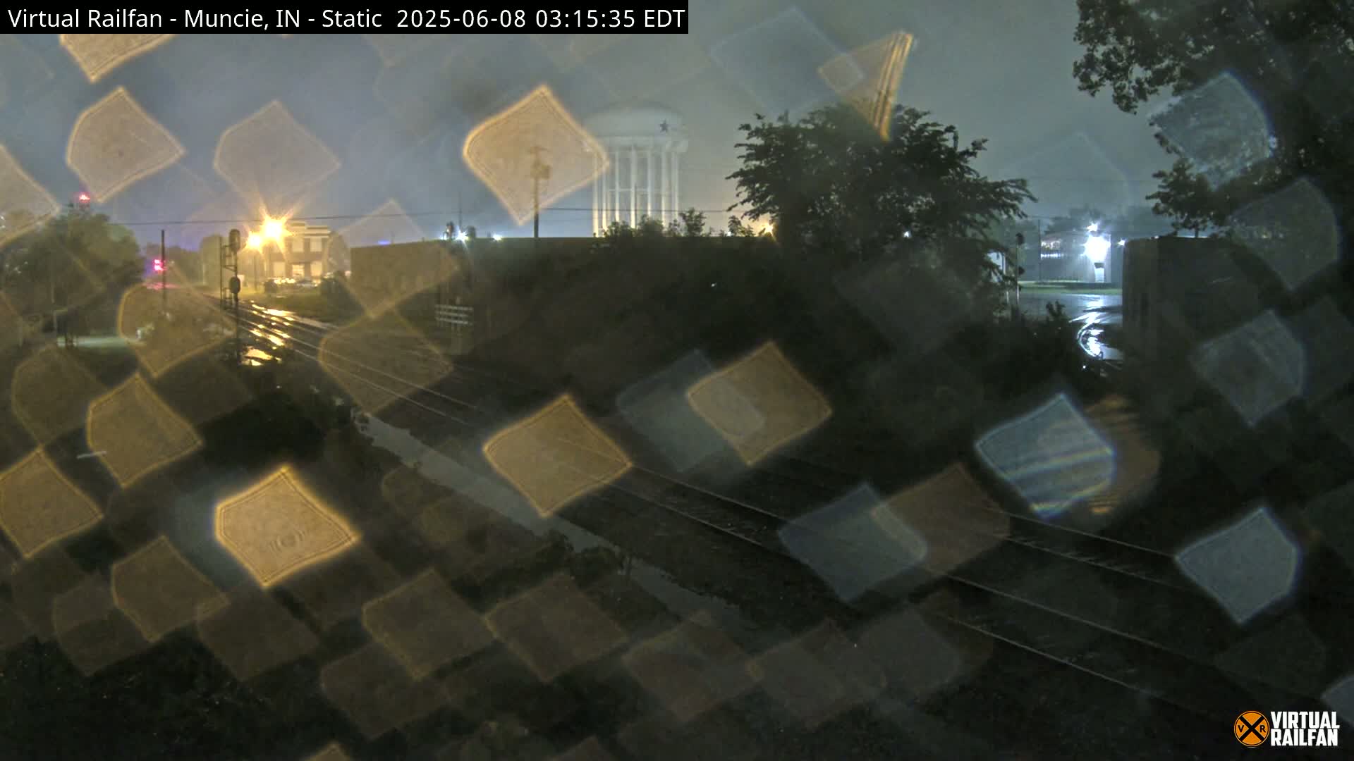 Rain falls at night on a railroad crossing near a water tower and buildings, with streetlights illuminating the wet streets.