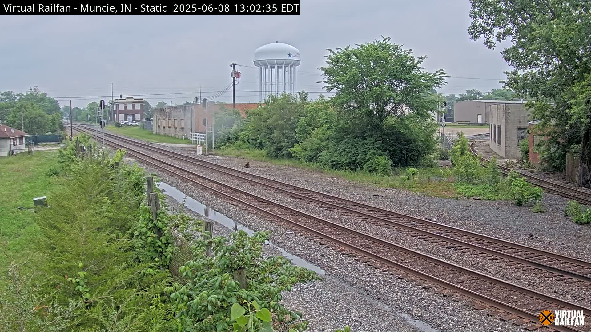 Two sets of train tracks stretch into the distance under an overcast sky, past industrial buildings and a water tower, with lush green vegetation along the sides.